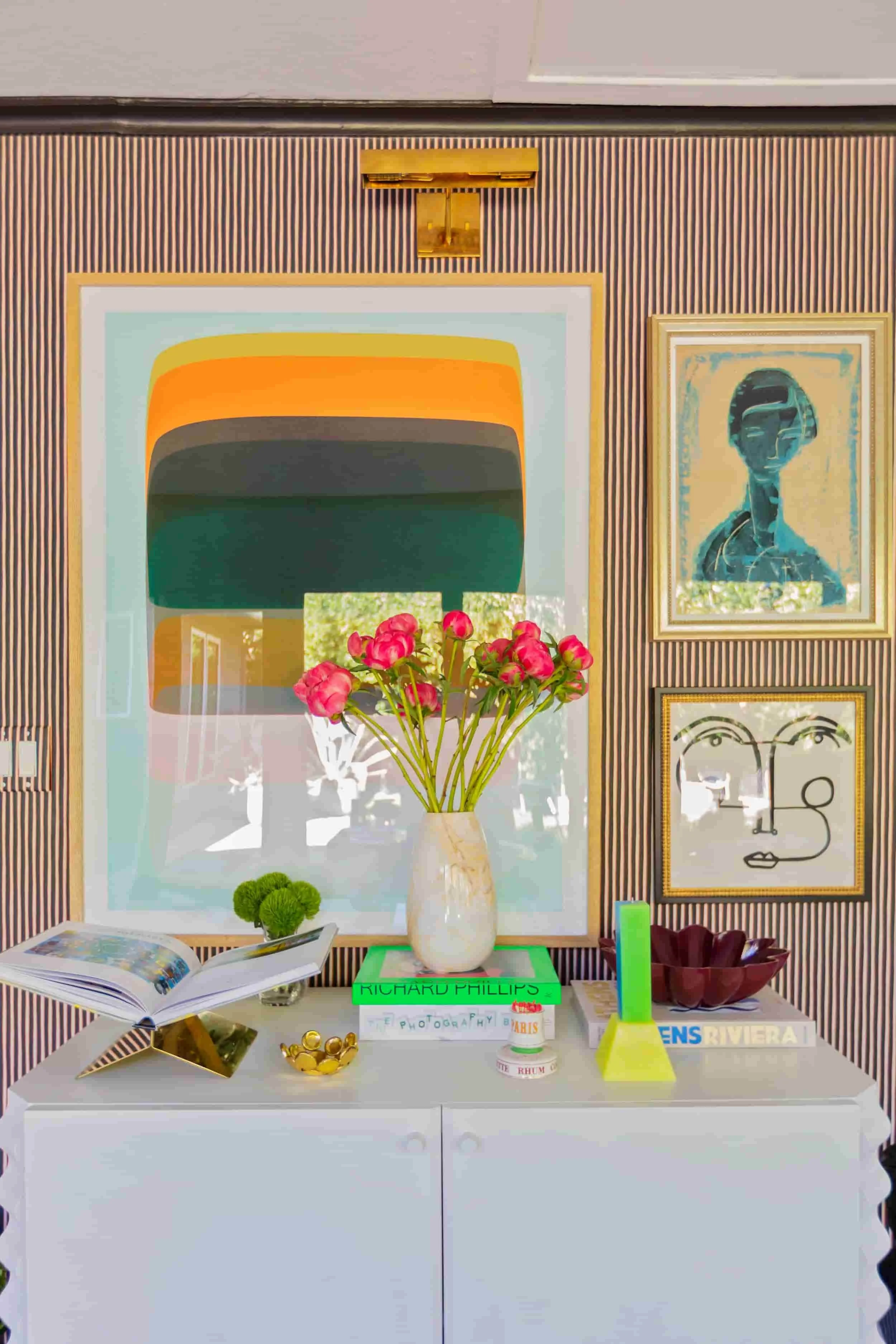 Decorative display on a white table featuring a white vase with pink flowers, an opened book, a small tray of gold coins, a green book titled 'Richard Phillips,' a yellow and green decorative object, a container of French perfume, and a sign with the