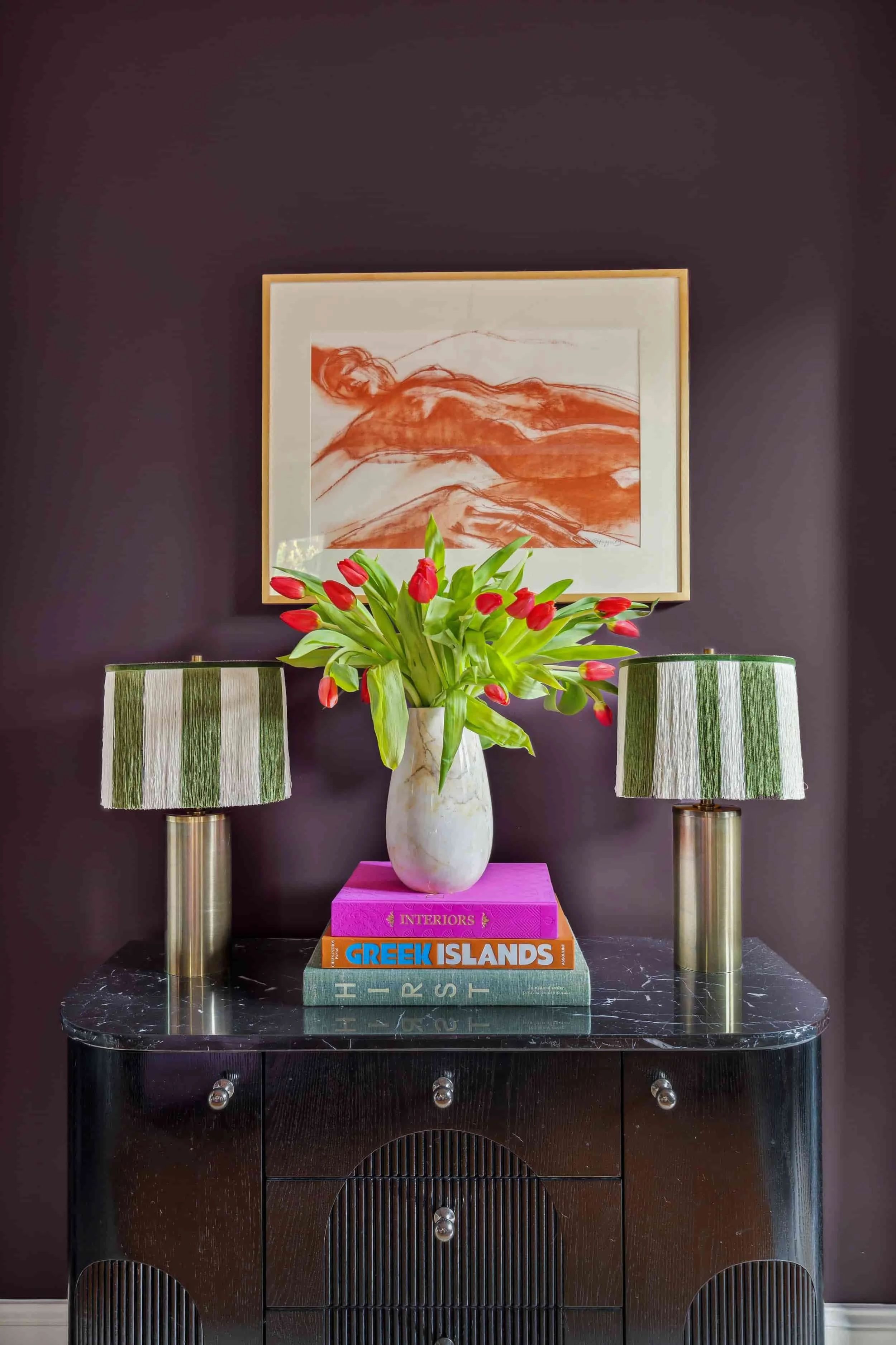 A black wooden cabinet with a marble top, decorated with a vase of red tulips in the center, two books underneath, and two matching table lamps on either side. A framed sketch of a reclining woman hangs on a dark wall behind.