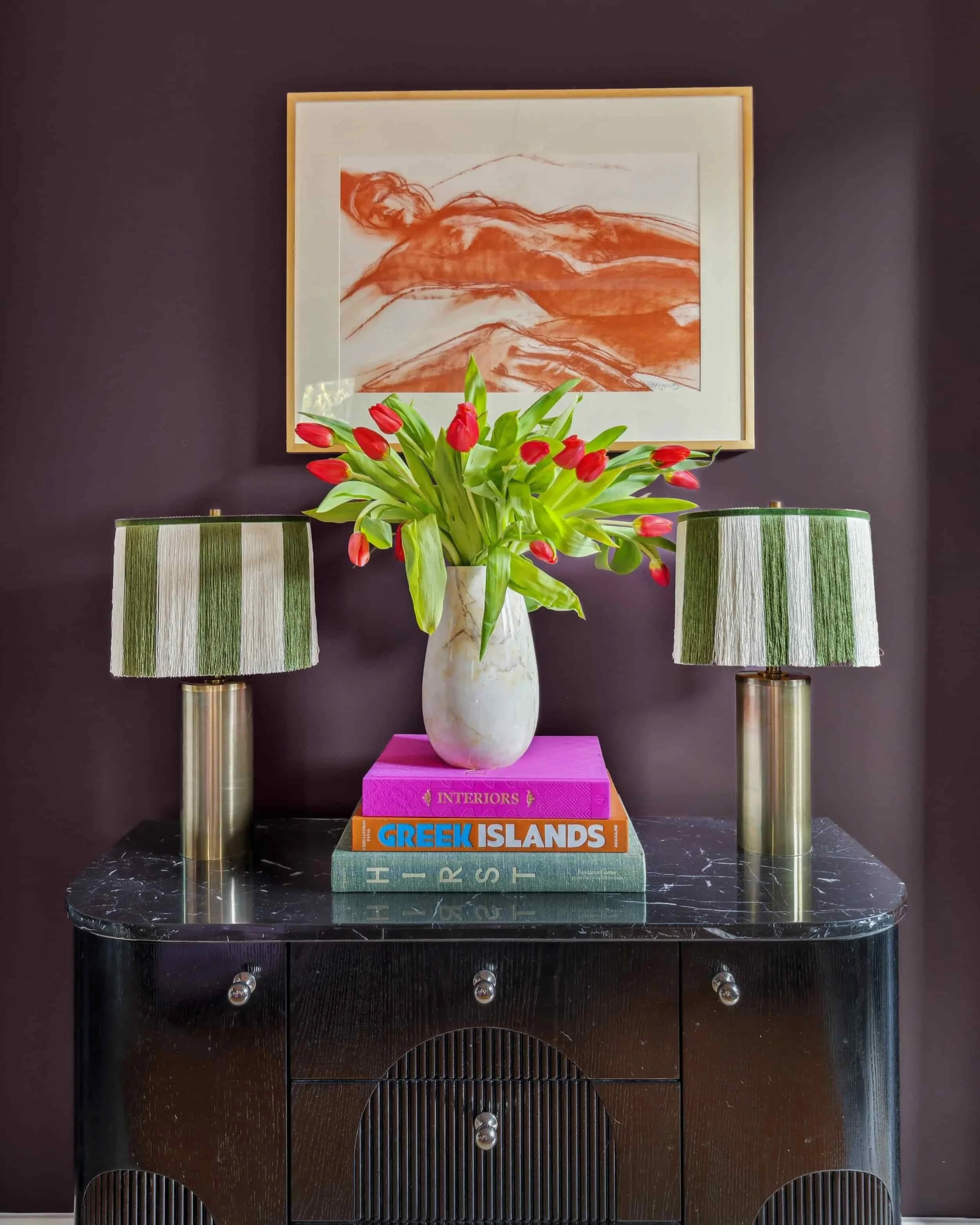 Black marble-top console table styled with green striped fringe lamps, red tulips in a marble vase and stacked coffee table books against a plum wall.