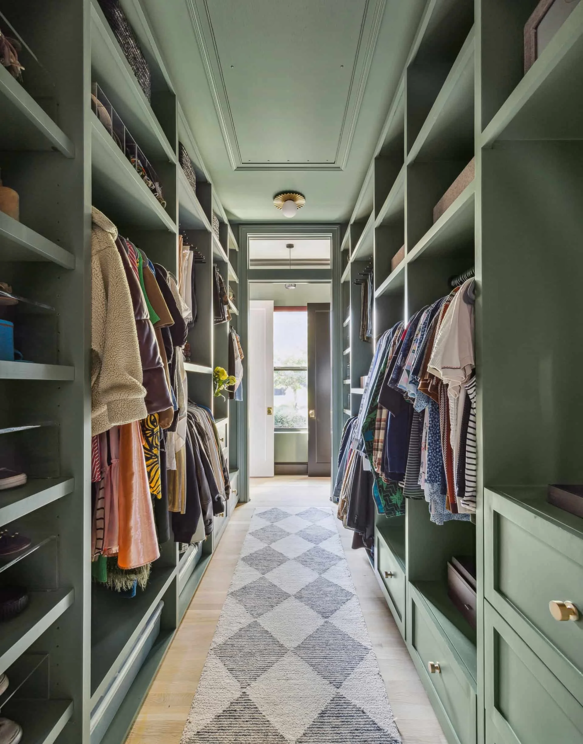 Walk-in closet designed by Hannah Goetz in Austin, Texas with sage green built-in storage, hanging clothes, and a patterned runner