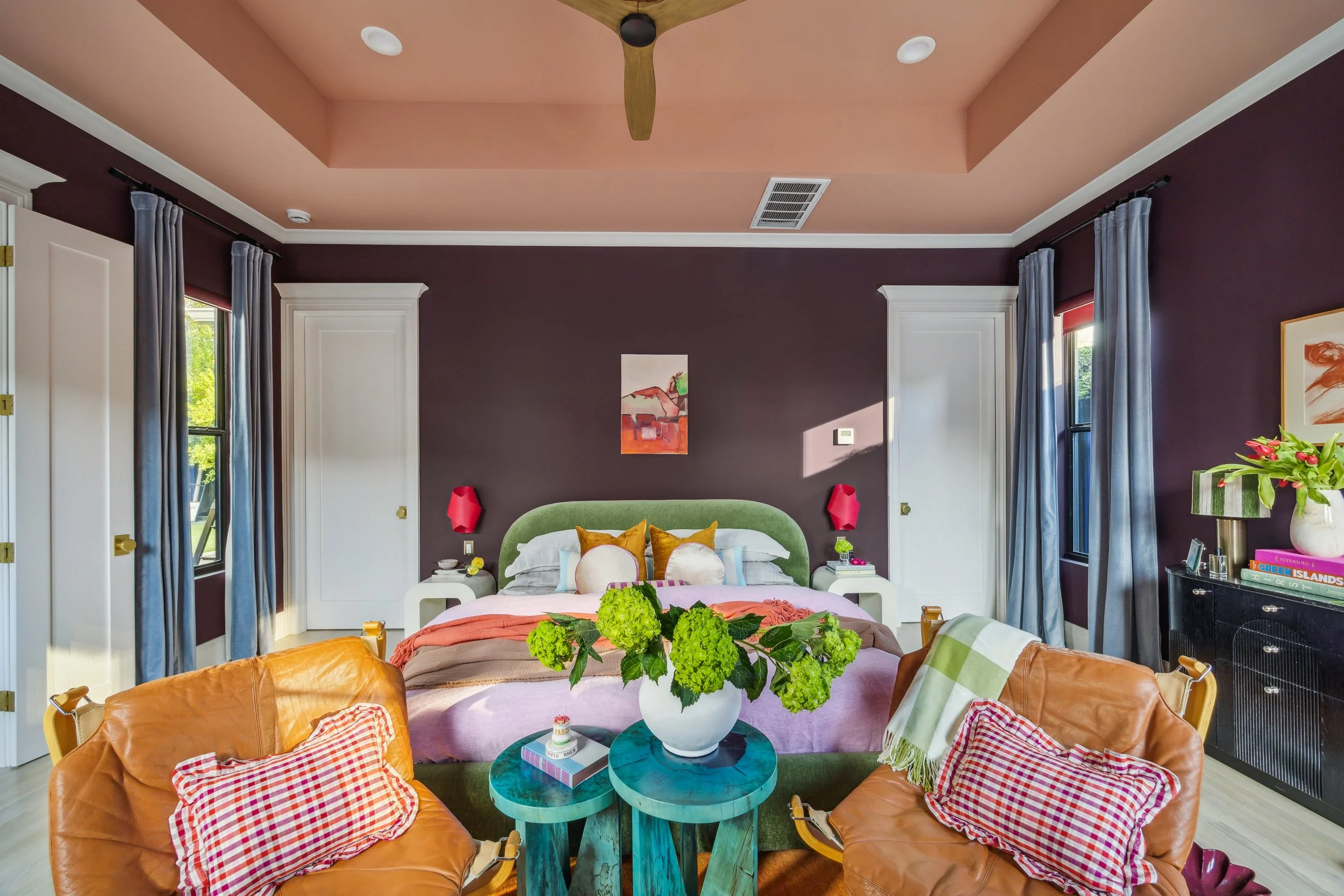Living room with purple walls, a bed with multiple pillows and blankets, a green upholstered headboard, and various chairs and furniture. Large windows with curtains, a ceiling fan, and artwork on the walls.