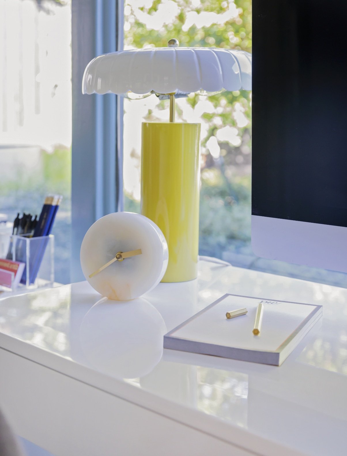 Desk styled by Hannah Goetz with a yellow table lamp, white clock, and notepad beside a window.