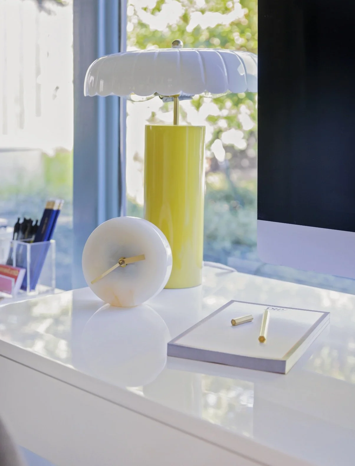 Desk styled by Hannah Goetz with a yellow table lamp, white clock, and notepad beside a window.