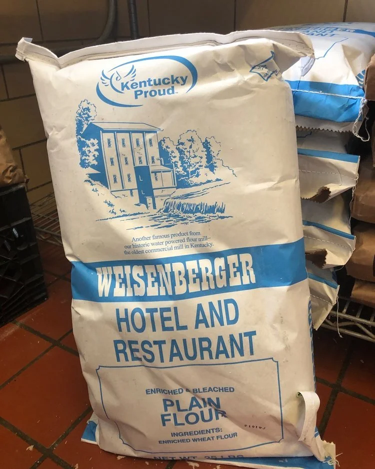 White and blue bag of Weisenberger Hotel and Restaurant plain flour, with a mountain and watermill illustration.