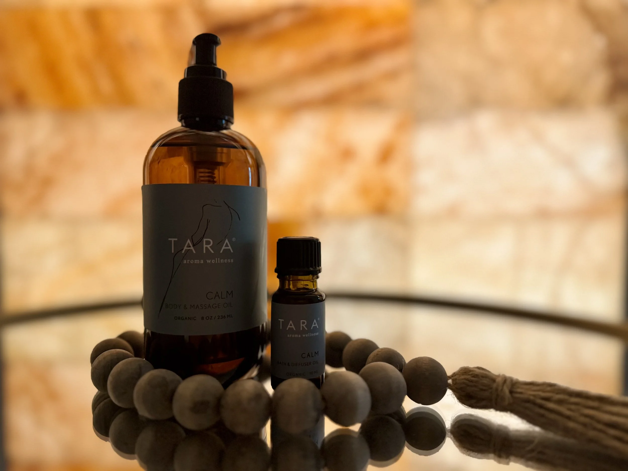 Two bottles of TARA aroma wellness essential oils, labeled 'CALM,' are placed on a reflective surface surrounded by round wooden beads and a tassel. The background is a blurred brick wall.