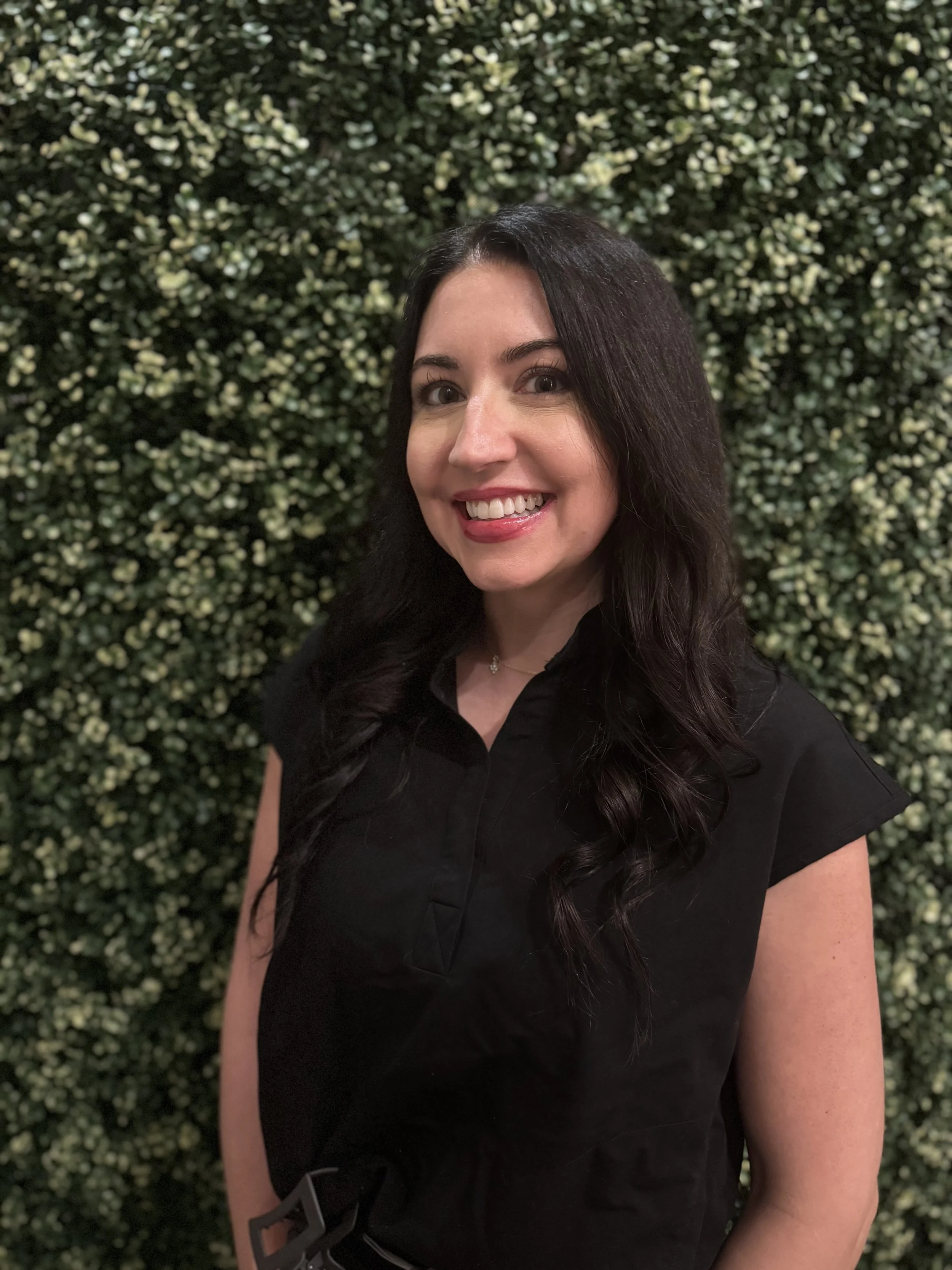 A woman with long dark hair taking a selfie in front of a dense, green leafy hedge. She is smiling and wearing a black shirt.
