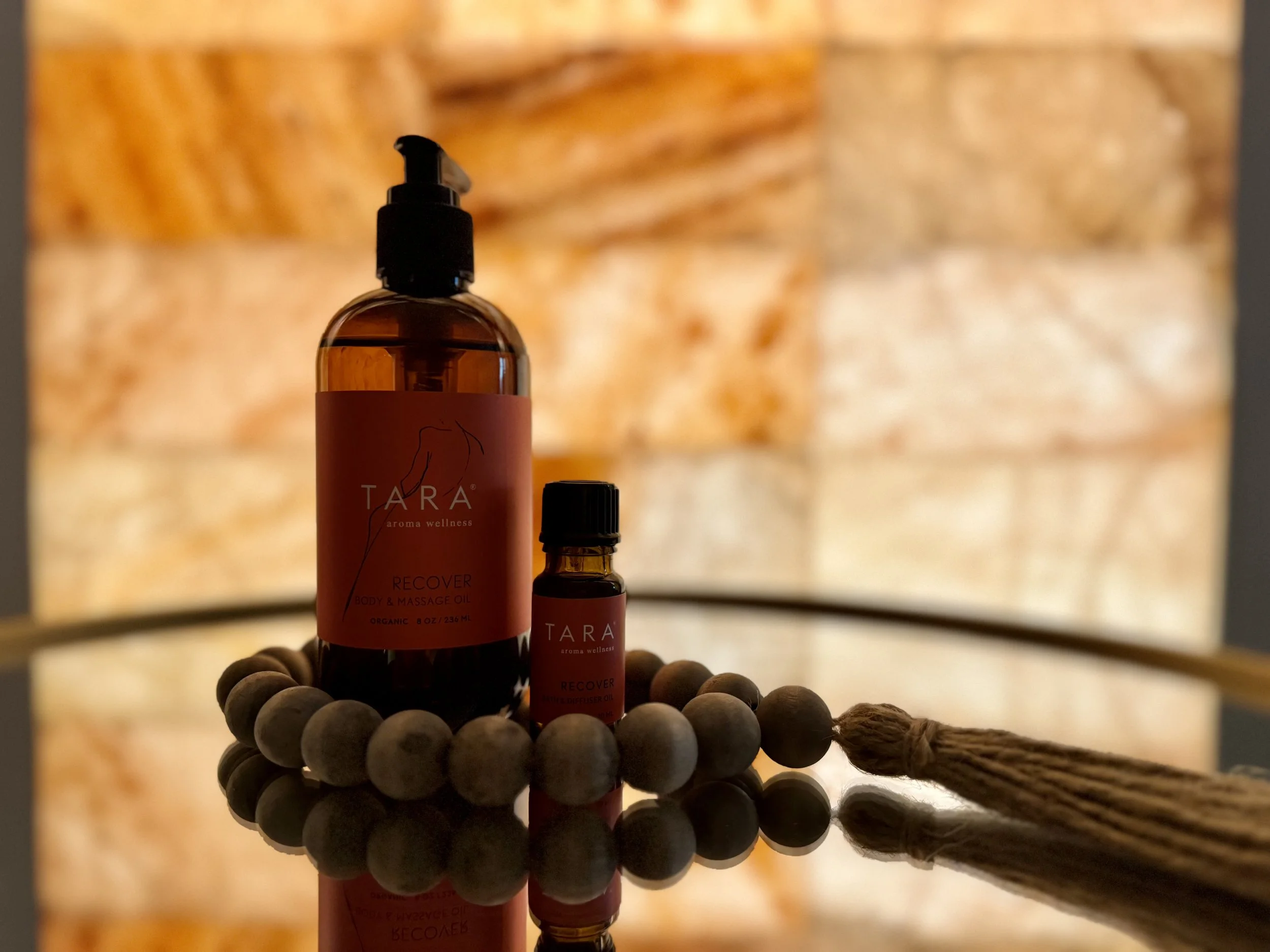 Two bottles of Tara aroma wellness product, one large and one small, placed on a reflective surface surrounded by a wooden bead bracelet and a brown tassel, with a blurred warm-toned brick wall background.
