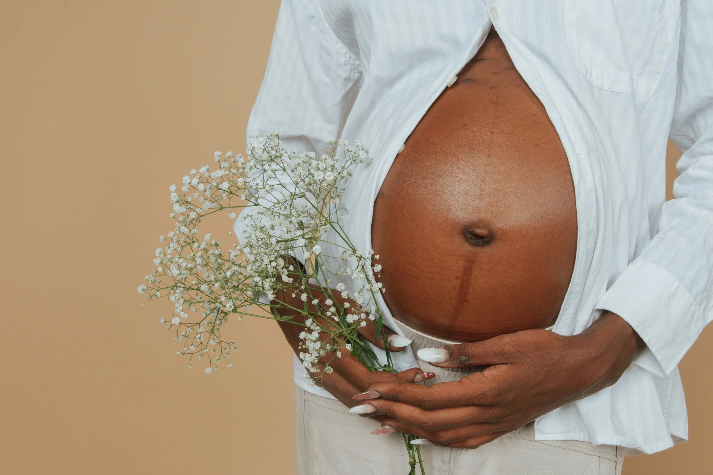 A person holding a bouquet of white flowers against a beige background, with bare pregnant belly visible.