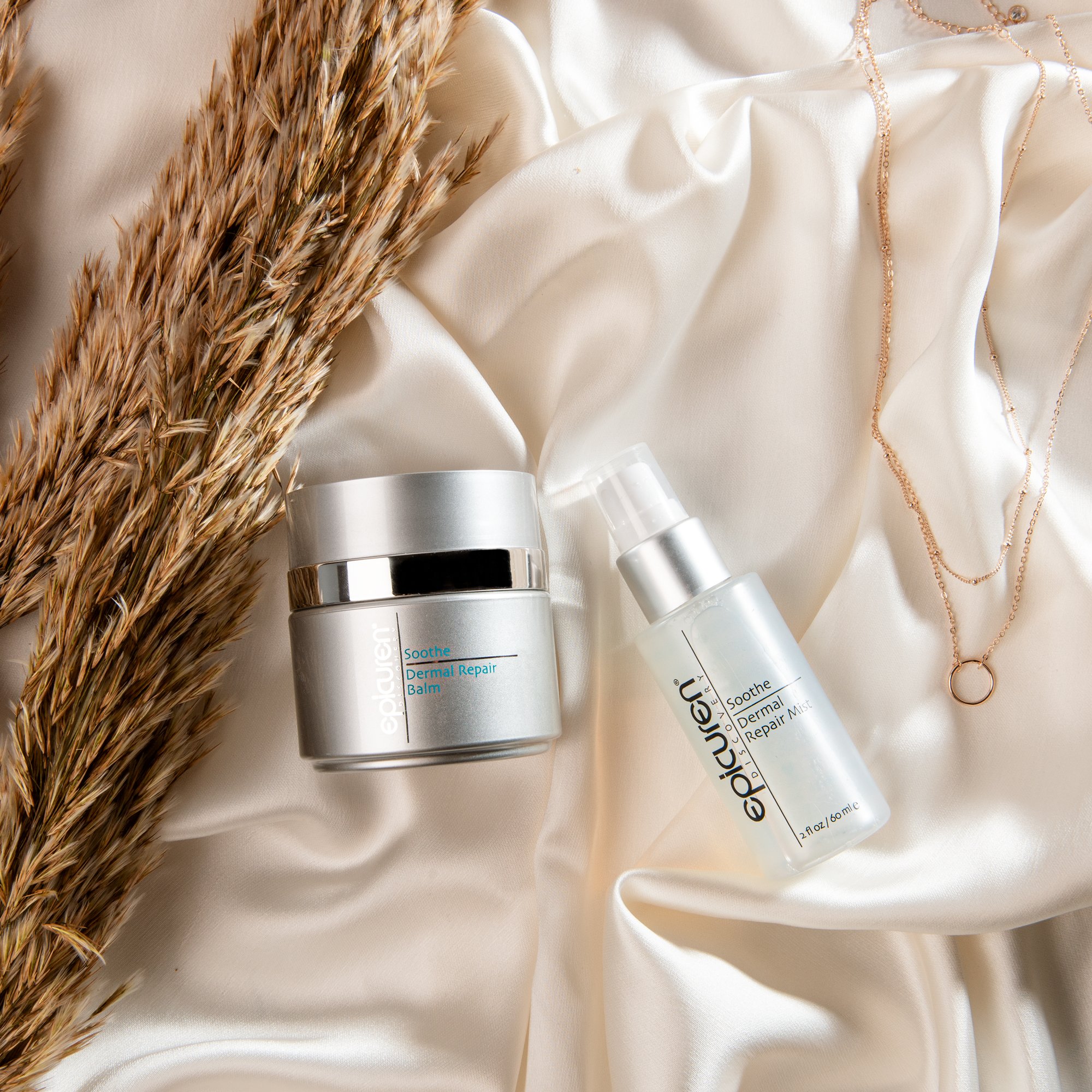 A jar of Epicuren Soothe Dermal Repair Balm and a spray bottle of Epicuren Soothe Dermal Repair Mist on cream-colored satin fabric, with dried wheat stalks and layered gold necklaces.