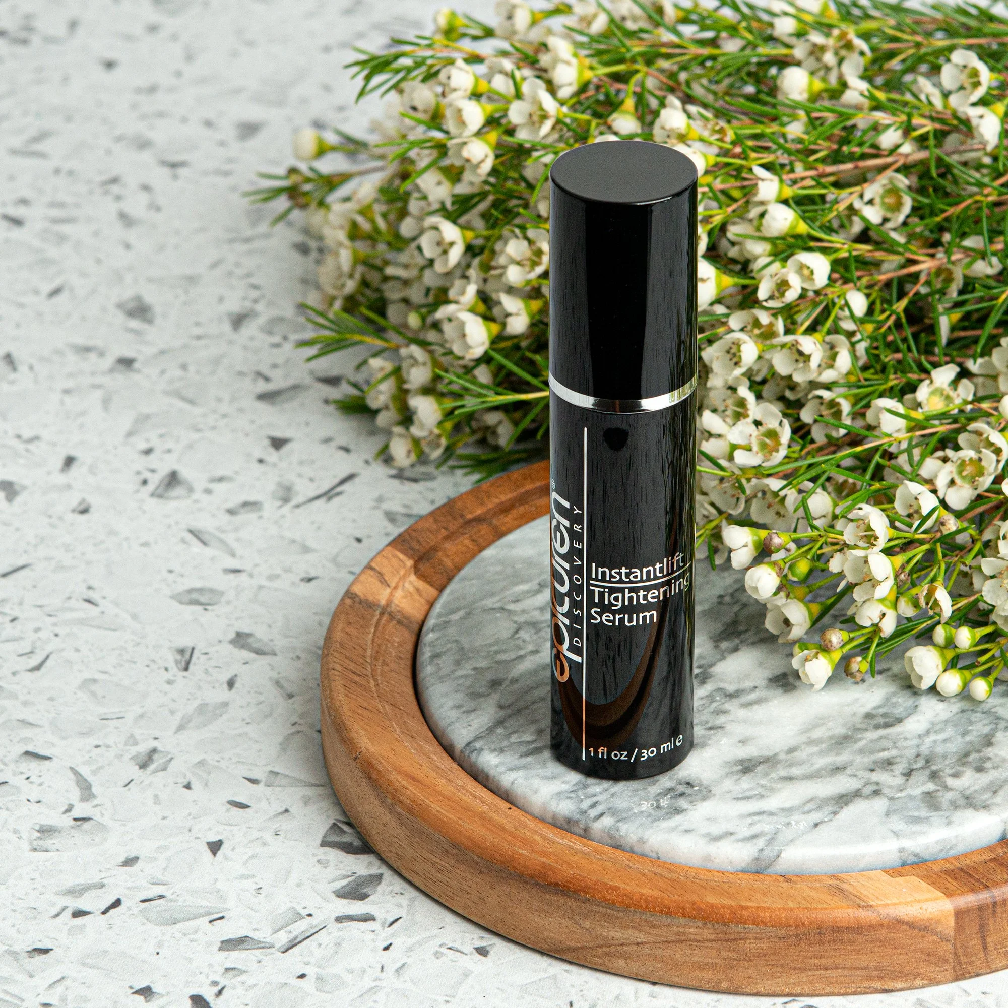 A black bottle of Glossier Instantlift Tightening Serum on a round marble tray, with a wooden border, surrounded by white flowers and green foliage on a speckled surface.