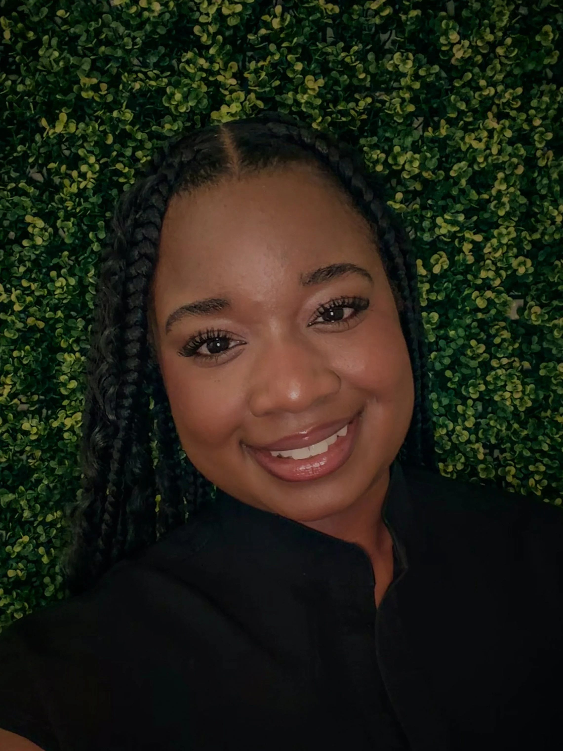A woman with braided hair, wearing a black top, smiling in front of a green leafy wall.