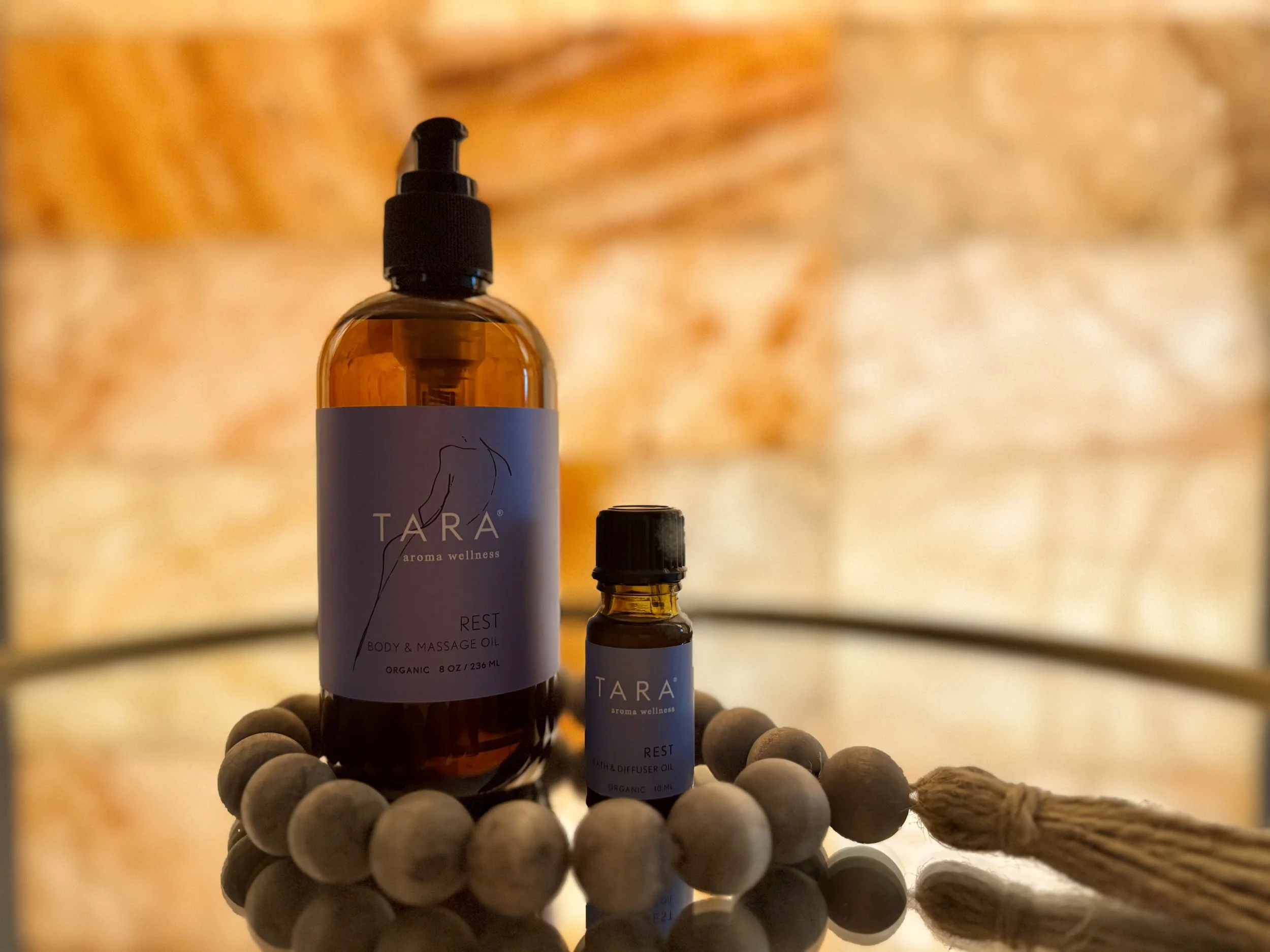 Two amber bottles labeled TARA aroma wellness, one larger and one smaller, placed on a reflective surface with a string of gray beads and a tassel in the foreground, against a warm, textured background.