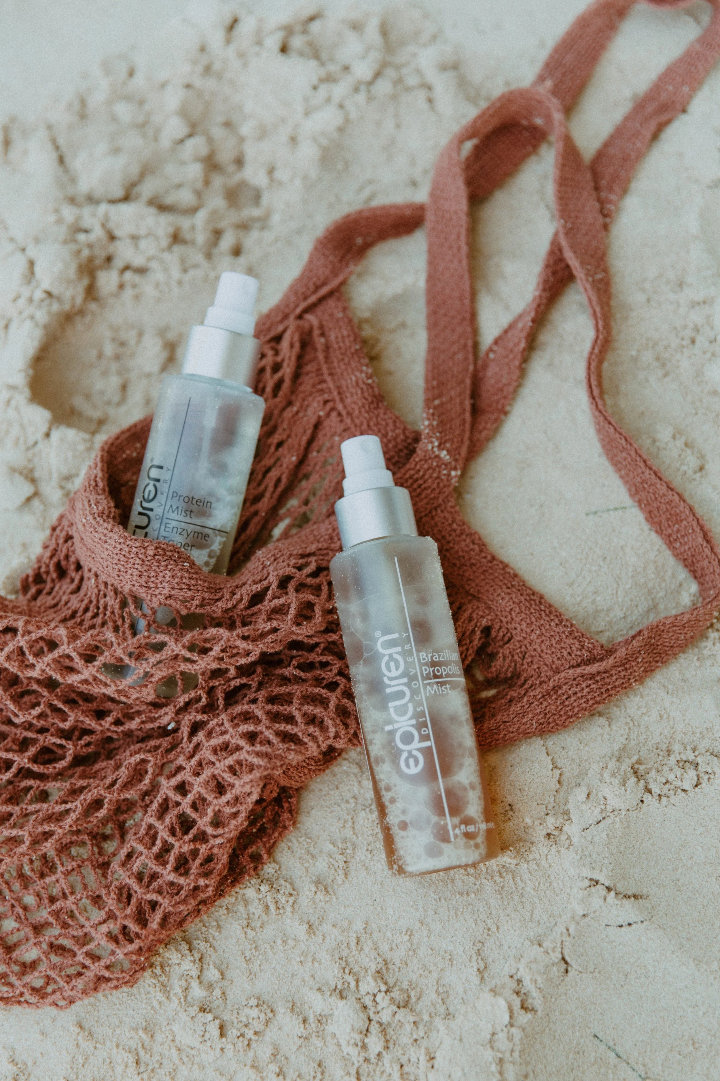 Two bottles of disinfectant mist placed on sandy ground inside a pink woven bag, with one bottle partially inside the bag and the other lying outside.