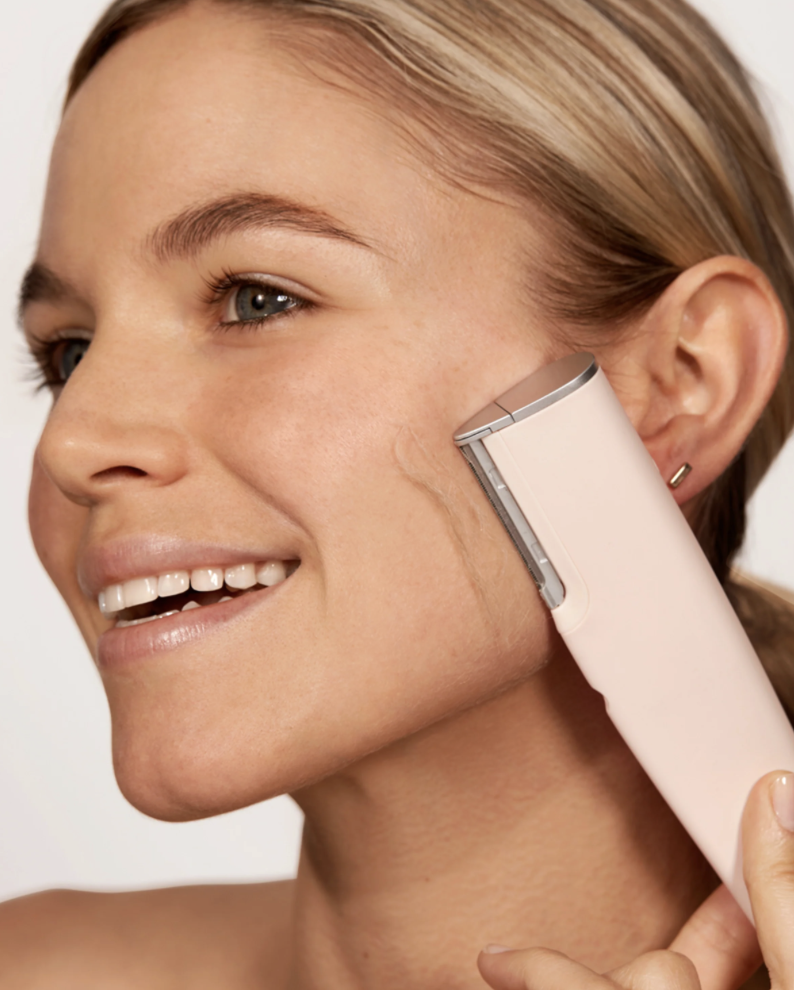A woman using an electric shaver to trim her sideburns, smiling and looking to the side.