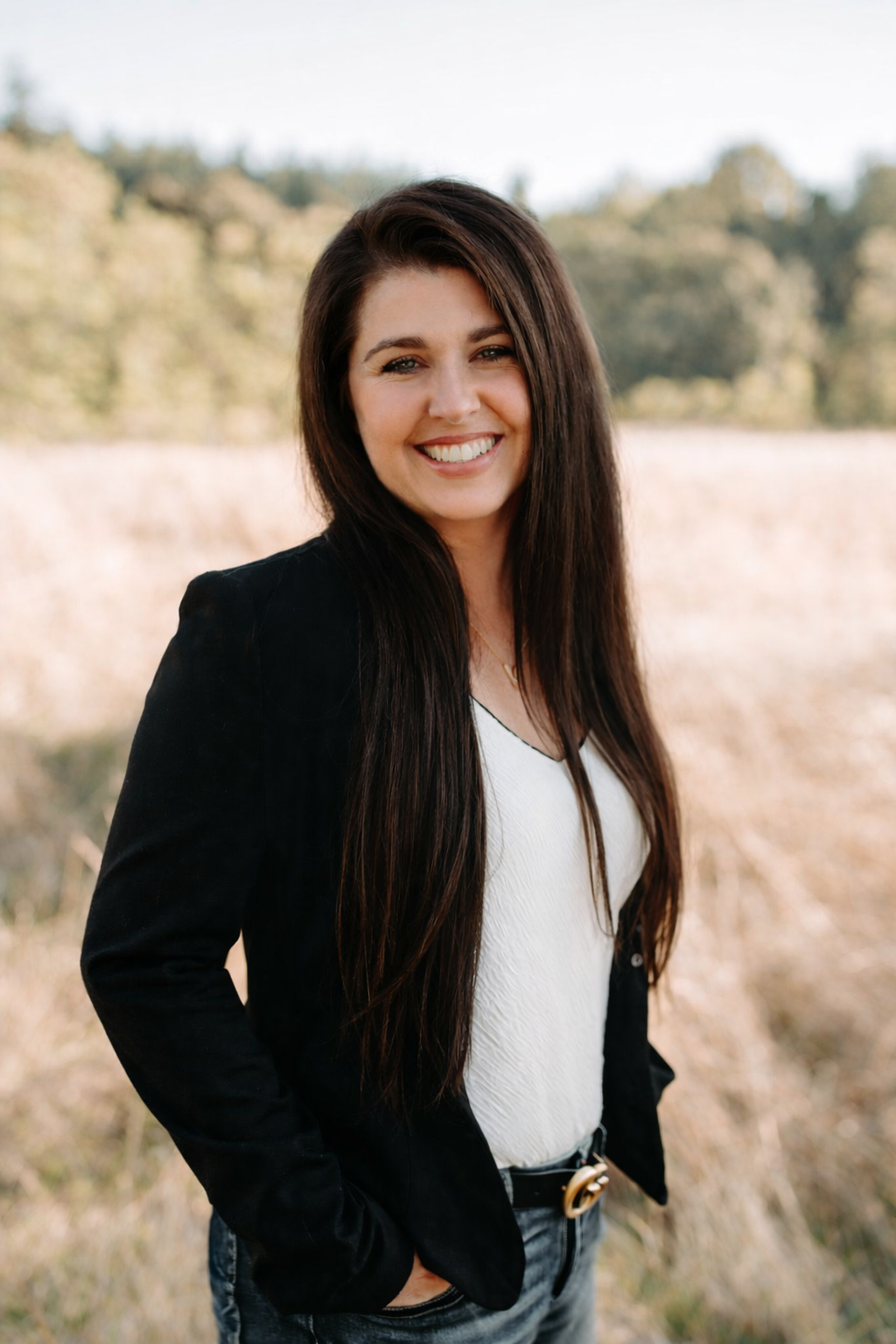 A woman with long brown hair smiling in front of a lush green leafy wall.