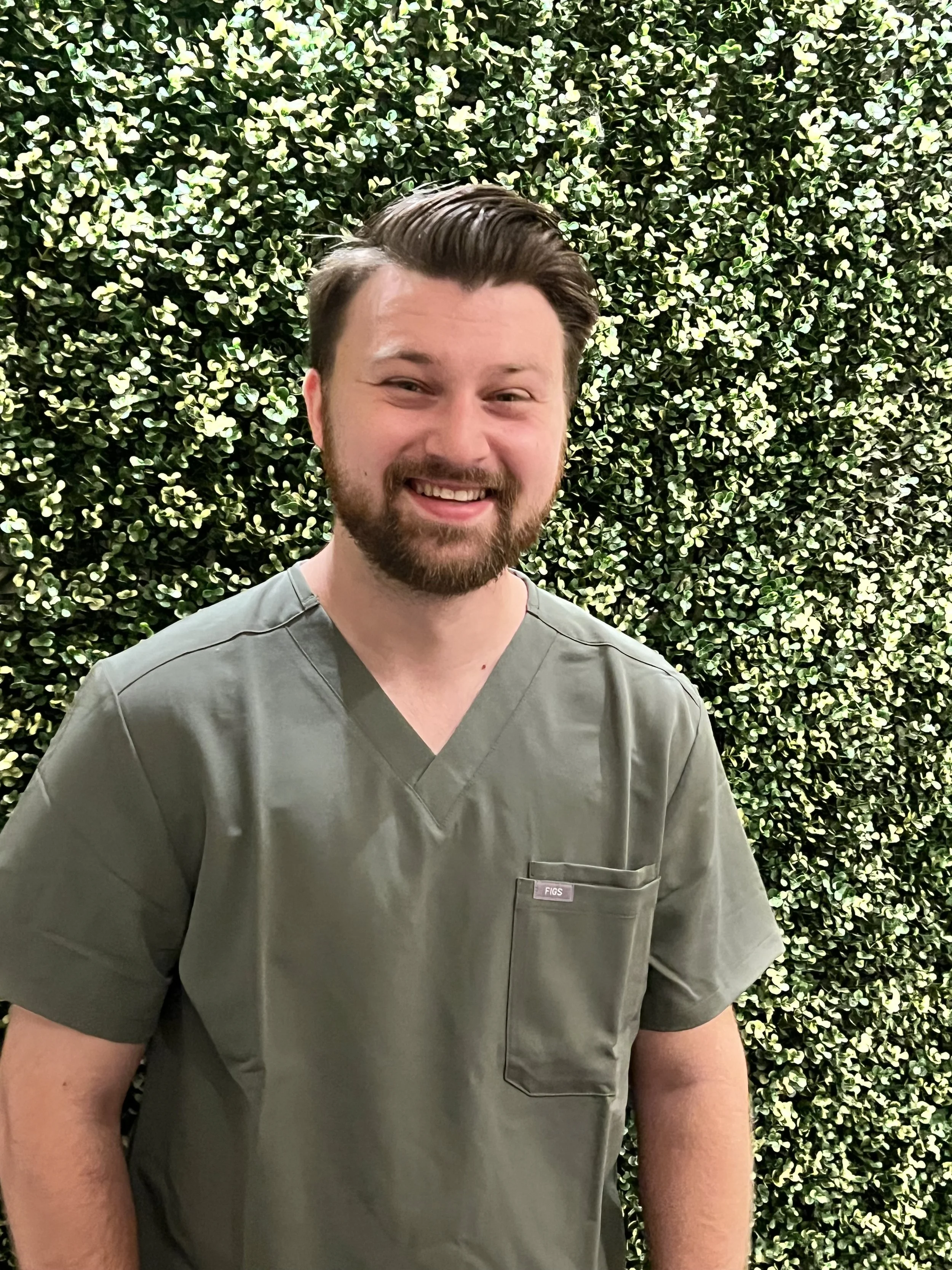 A young man with brown hair and a beard wearing green medical scrubs standing in front of a leafy green background.