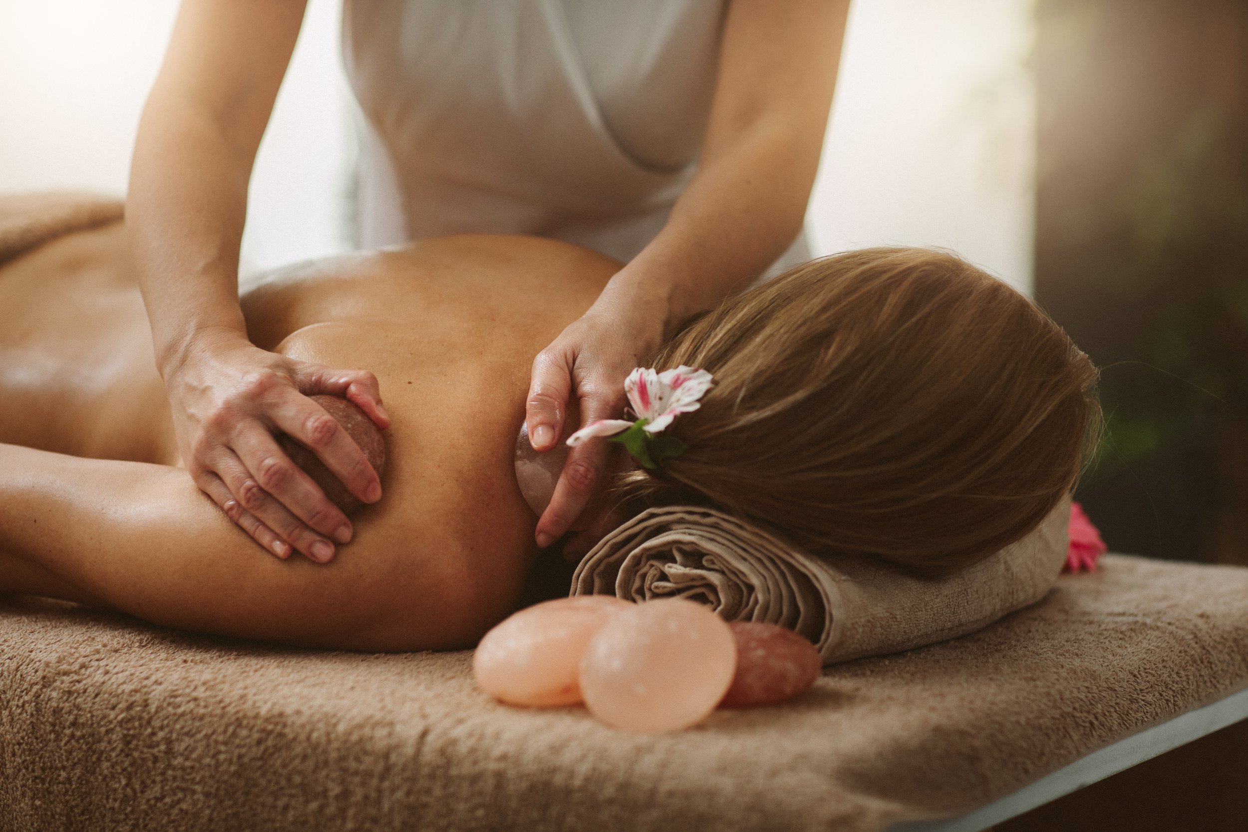 A woman is receiving a massage in a spa setting, lying face down on a massage table. The massage therapist is using their hands to apply pressure to her back. There is a decorative flower in her hair, and some polished stones are placed nearby on the table.