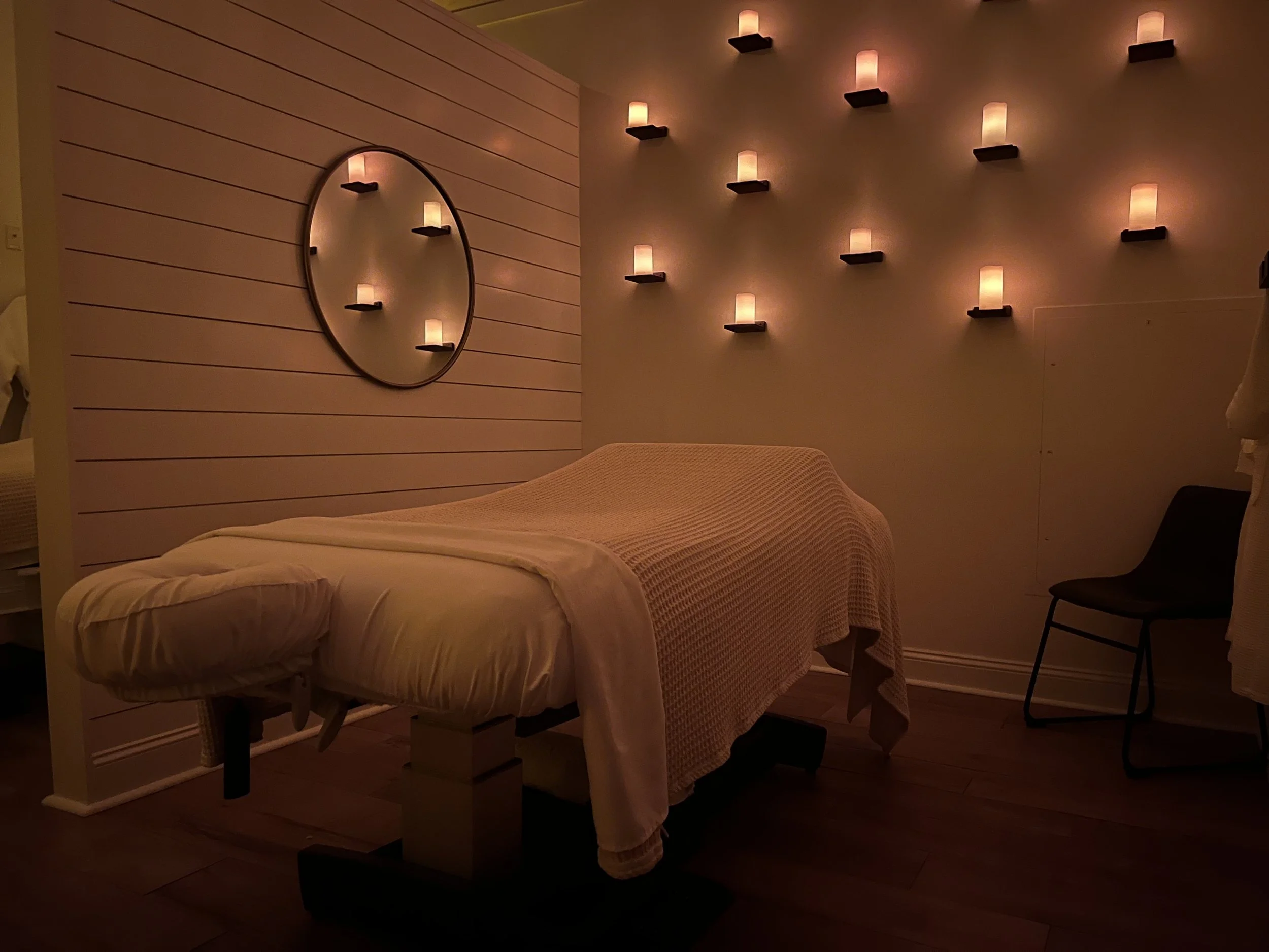 A massage room with a massage table draped in white linens, illuminated by warm candle lights on the wall, creating a relaxing ambiance.