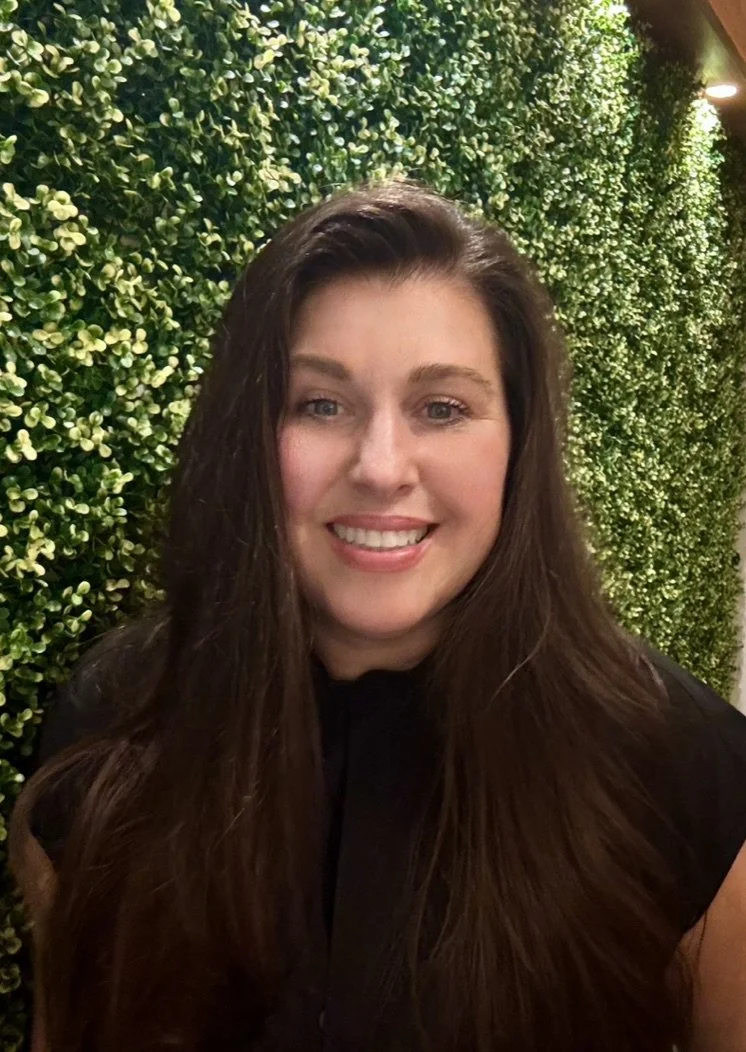 A woman with long brown hair smiling in front of a lush green leafy wall.