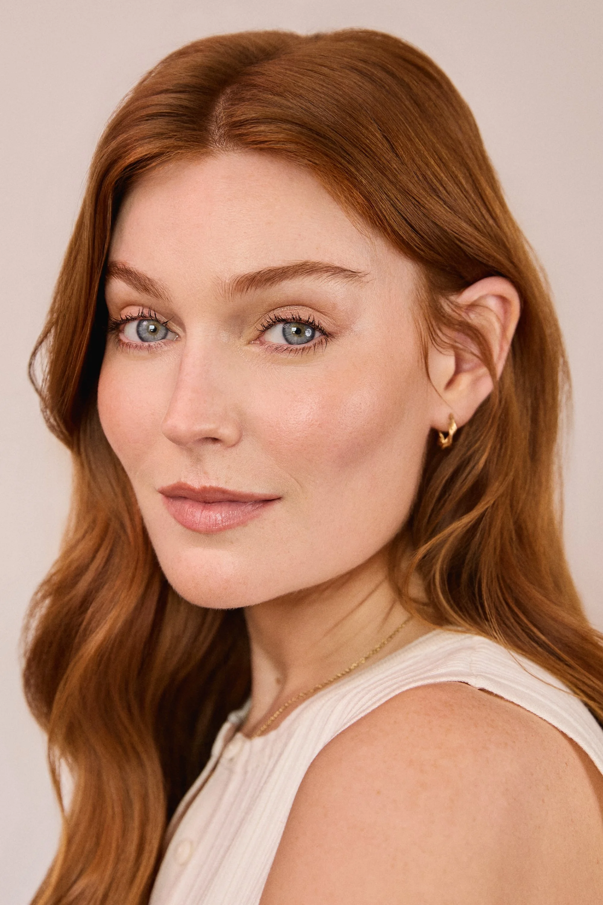 Close-up of a woman with red hair, blue eyes, and a light makeup look, wearing small gold earrings, a gold chain necklace, and a white sleeveless top.