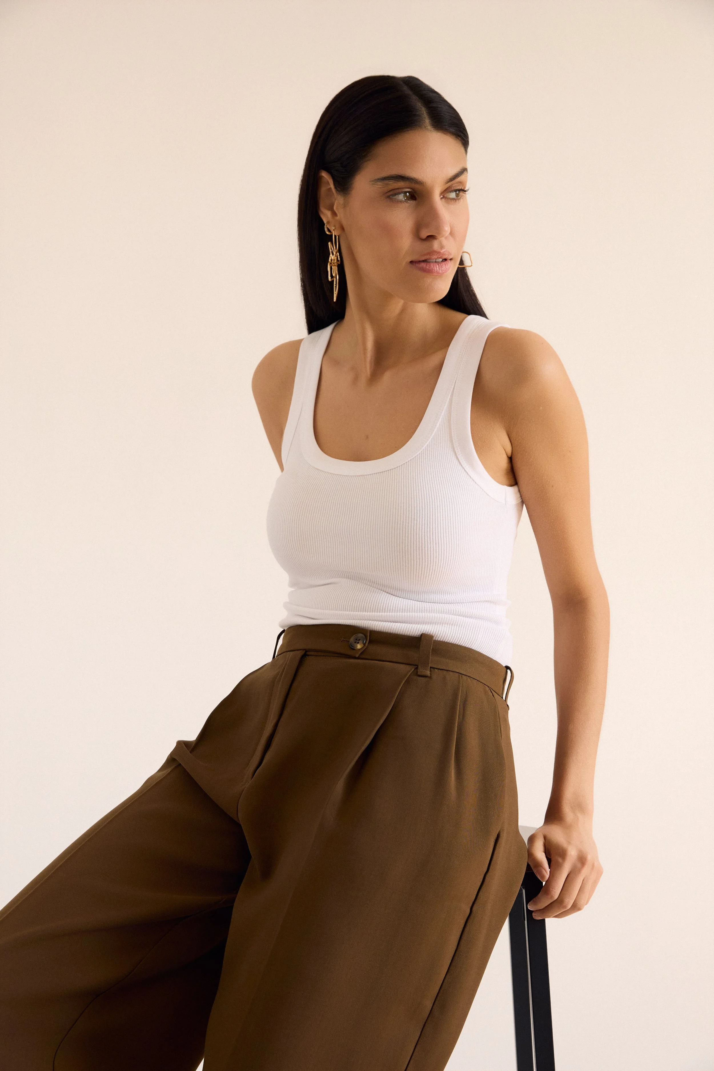 A woman with dark hair wearing a white tank top and brown high-waisted trousers, sitting on a chair against a plain white background.
