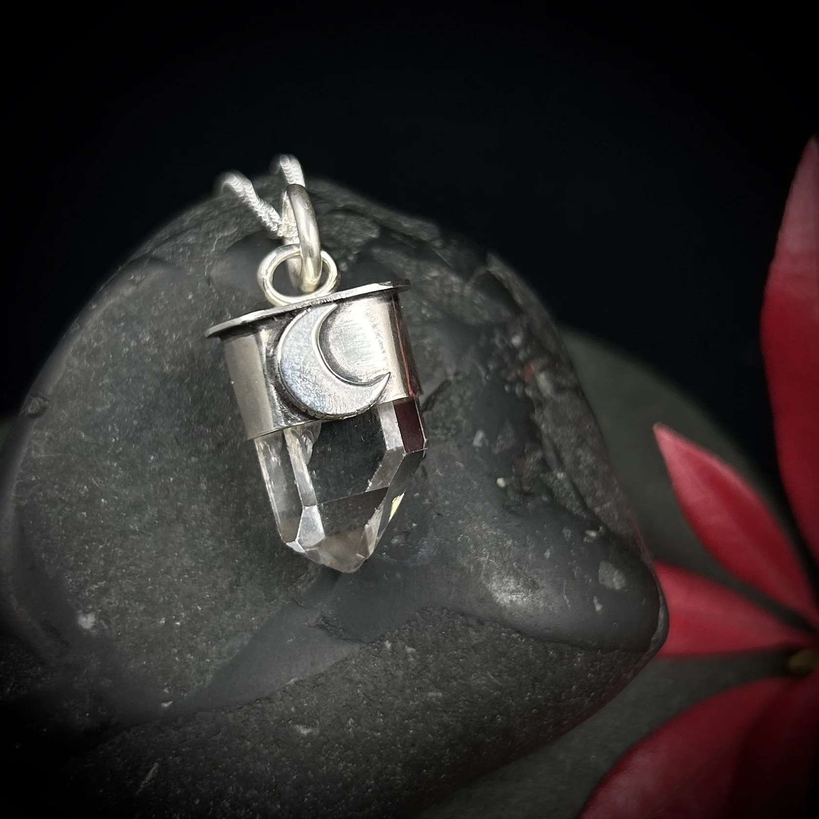 Small clear Tibetan quartz point set in silver with a large waning crescent moon design on a fine silver chain against a grey rock and bright red foliage