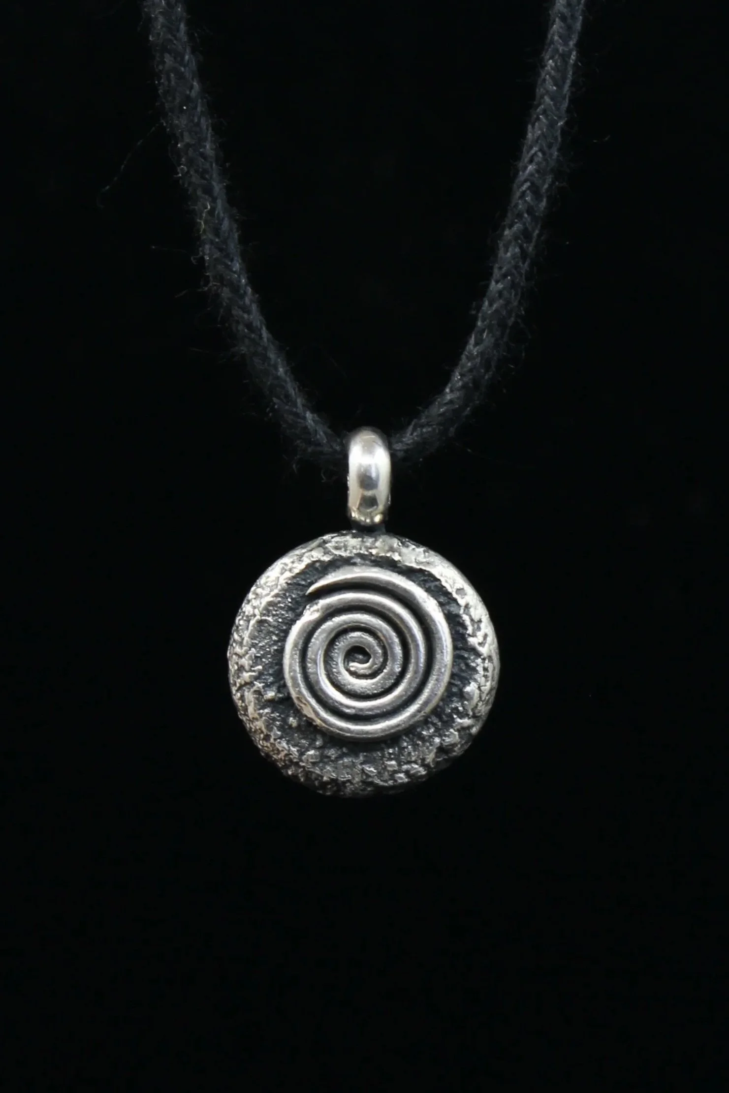Round and heavily textured silver pendant bearing a raised and eroded spiral design. The silver has been oxidised to match the black cord and is displayed with a black background. 