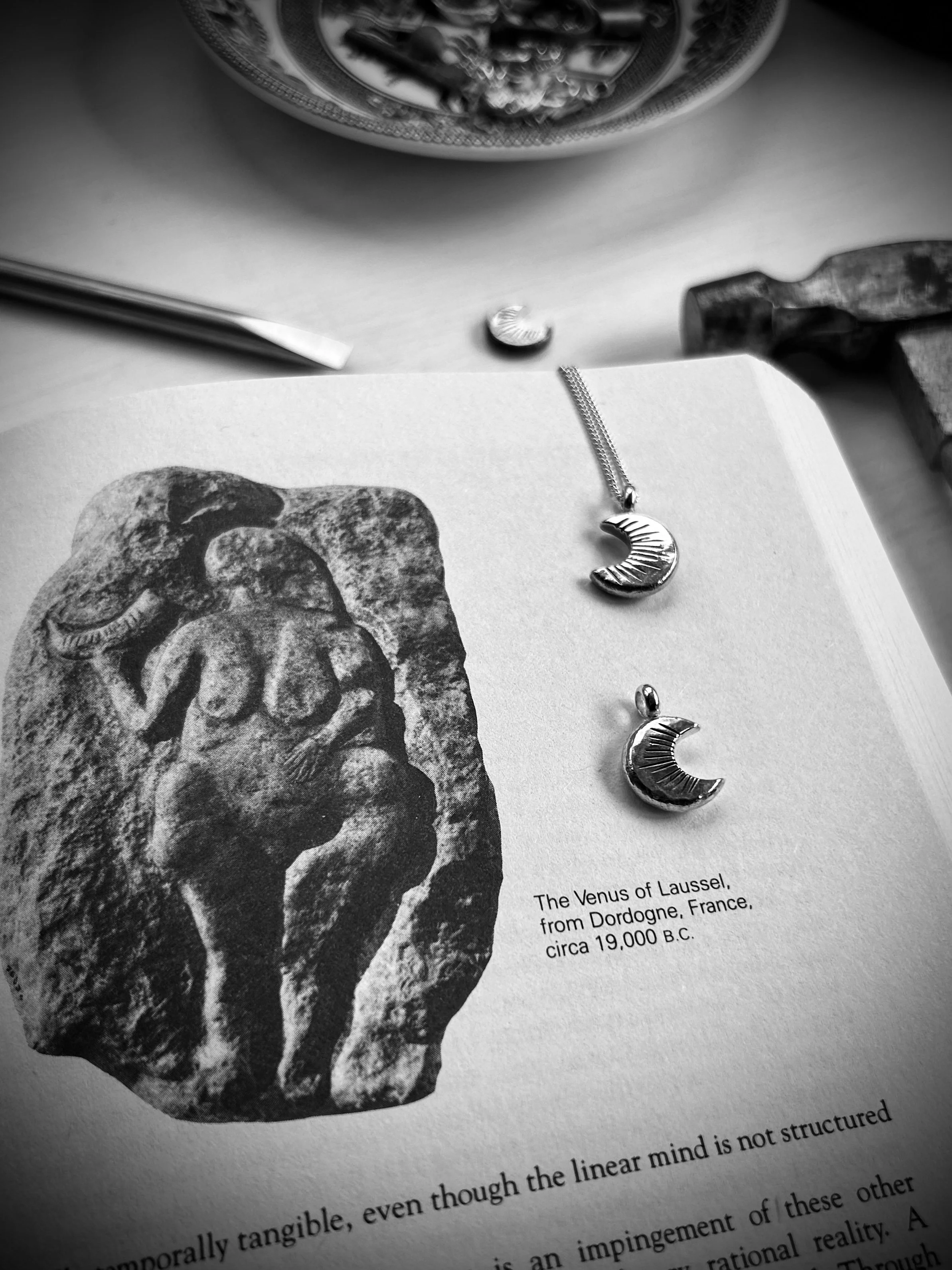 Two forged silver crescent moon pendants lying on an open book depicting Venus of Laussel. The thirteen notched marks are visible along with the jewellery tools, hammer and punch, that made them.