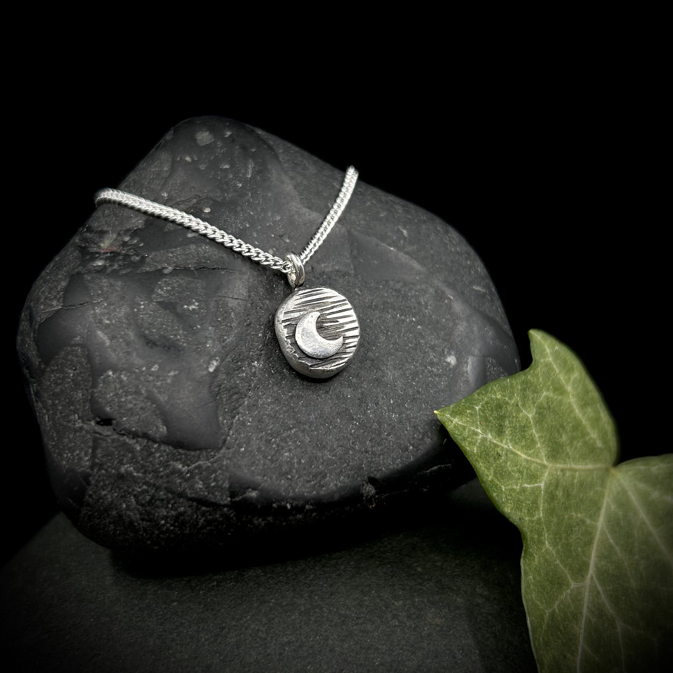Small circular solid silver pebble with silver crescent moon set on background of textured lines, on a fine silver chain against a grey rock