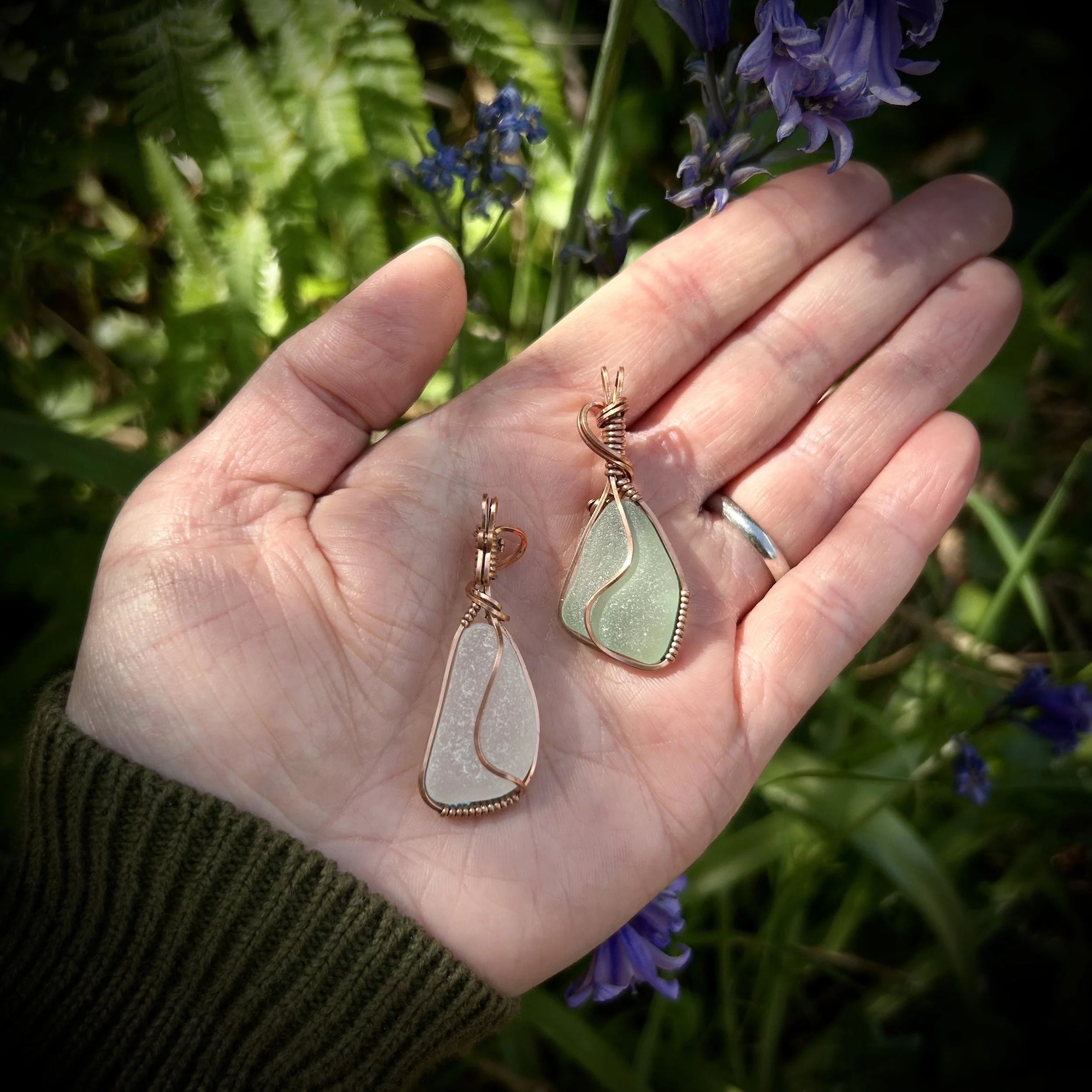 One clear and one green sea glass pendants wrapped in bright copper and lying on a women’s palm against a fern and bluebell backdrop.