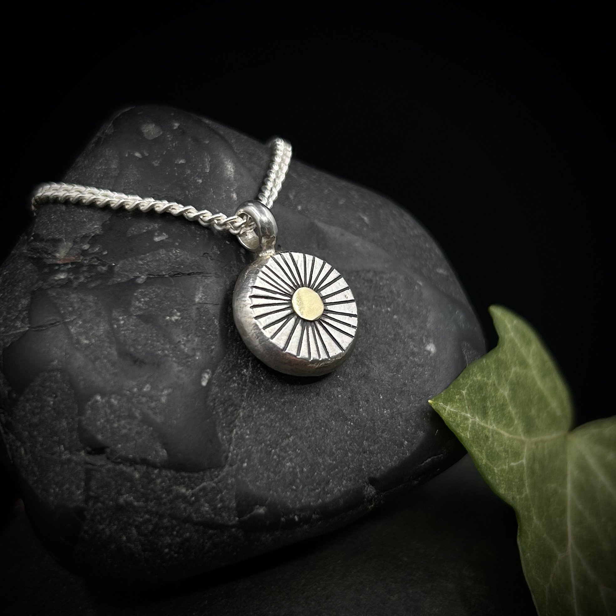 Side view showing the thickness of the solid silver sun pendant and close up of the textured lines radiating out from the 18k gold centre, positioned against a grey rock and green ivy leaf