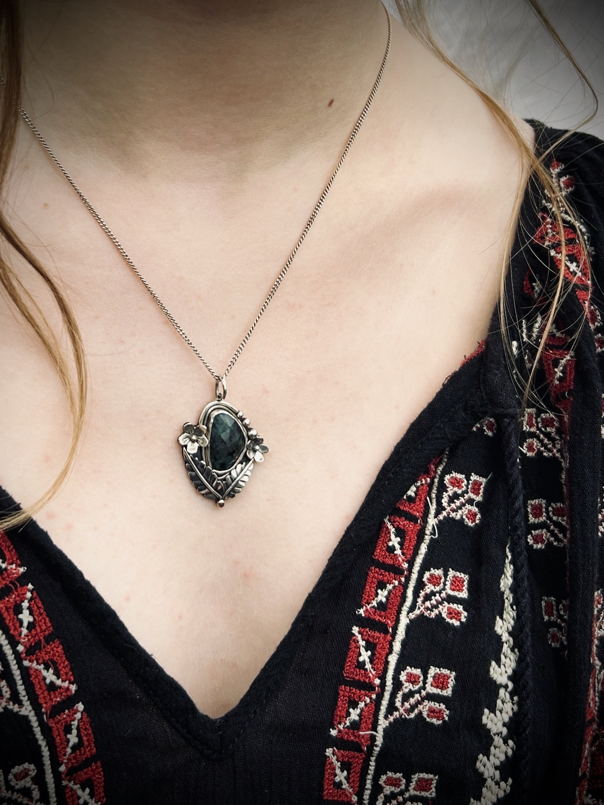 Model in black embroidered peasant blouse wearing silver and emerald floral pendant on a thin silver chain.