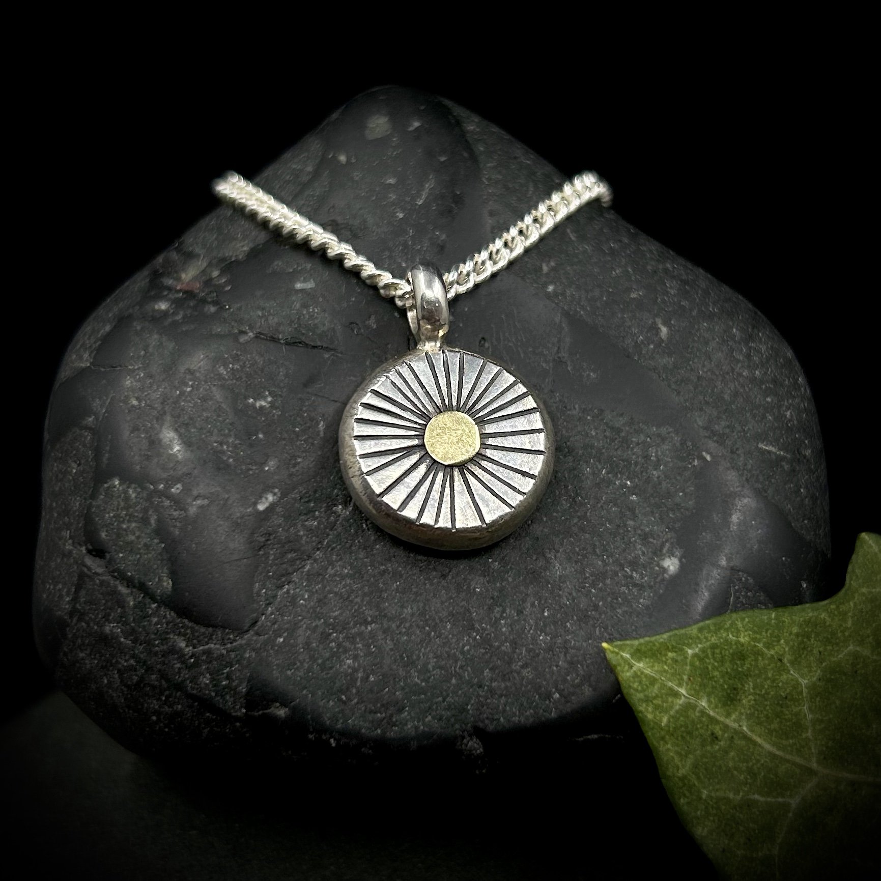 Solid silver sun pendant, a heavy circular pebble of silver with textured lines radiating out from an 18k gold centre, on a silver chain against grey rocks