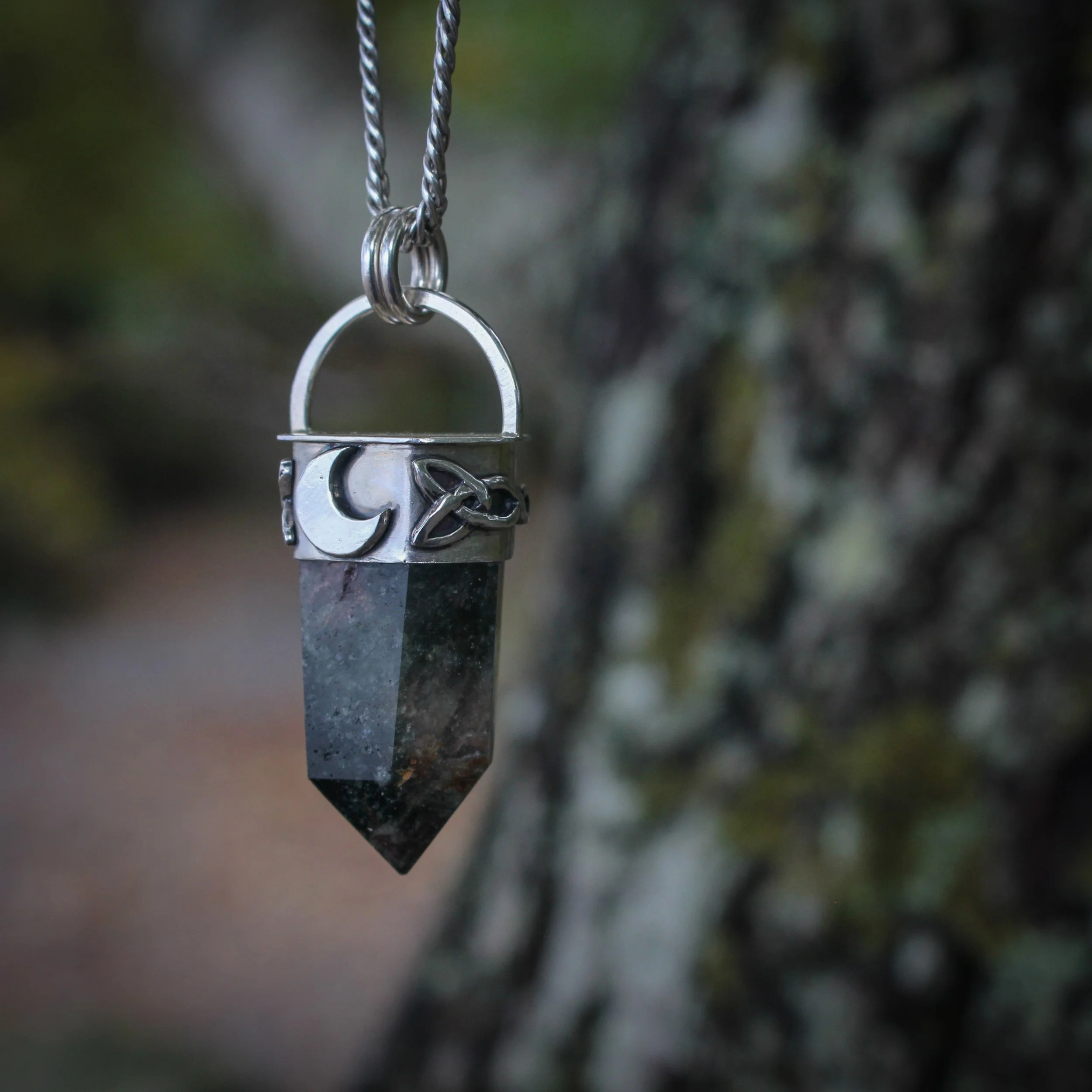 Dark coloured quartz crystal point set in silver with crescent moon and Celtic knotwork details hanging next to a dark tree