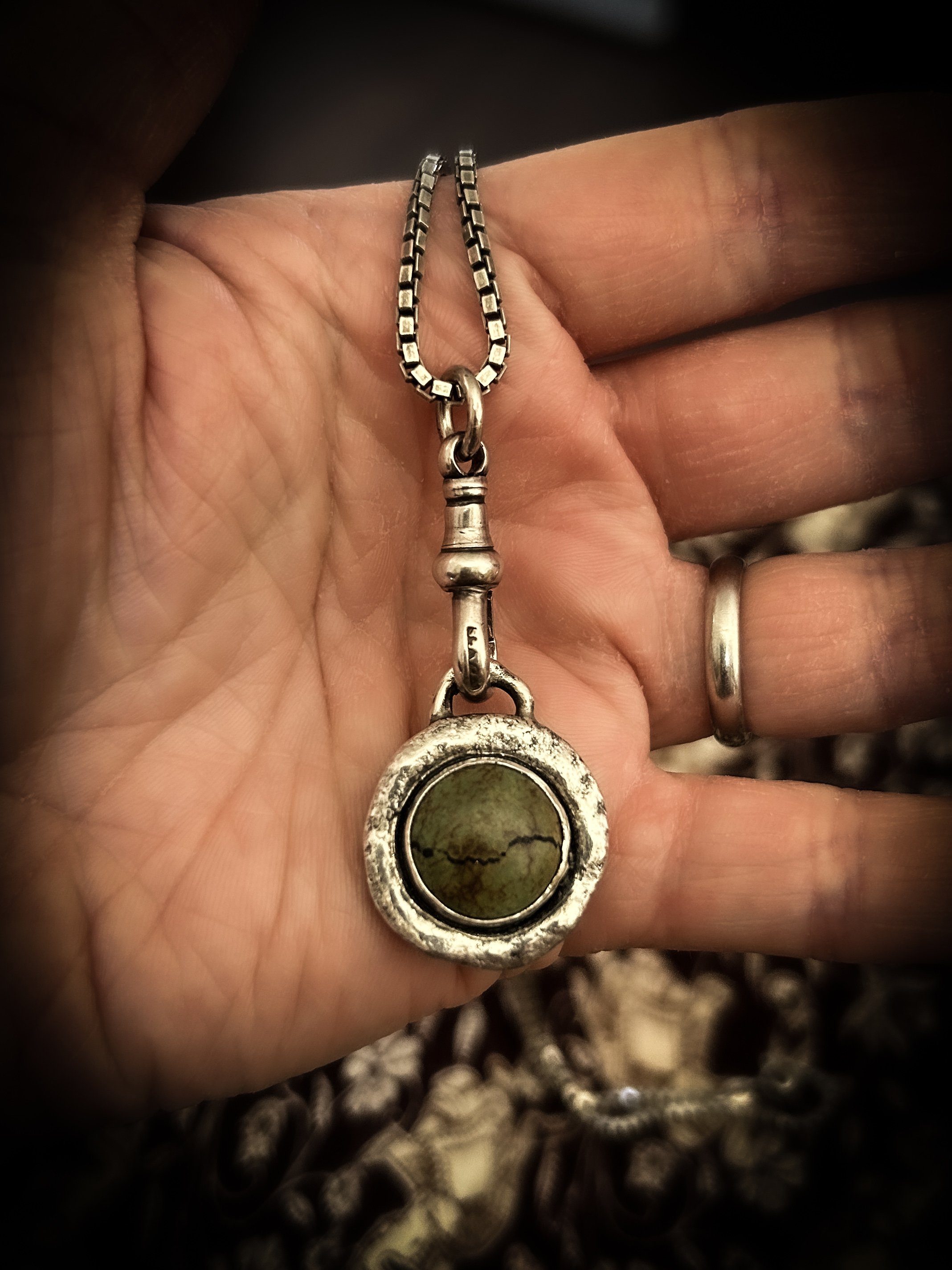 A large and sculptural round silver pendant lies in an open palm. The silver is organic and textured, in the middle is a green-brown earthy stone with a visible black crack across the surface.