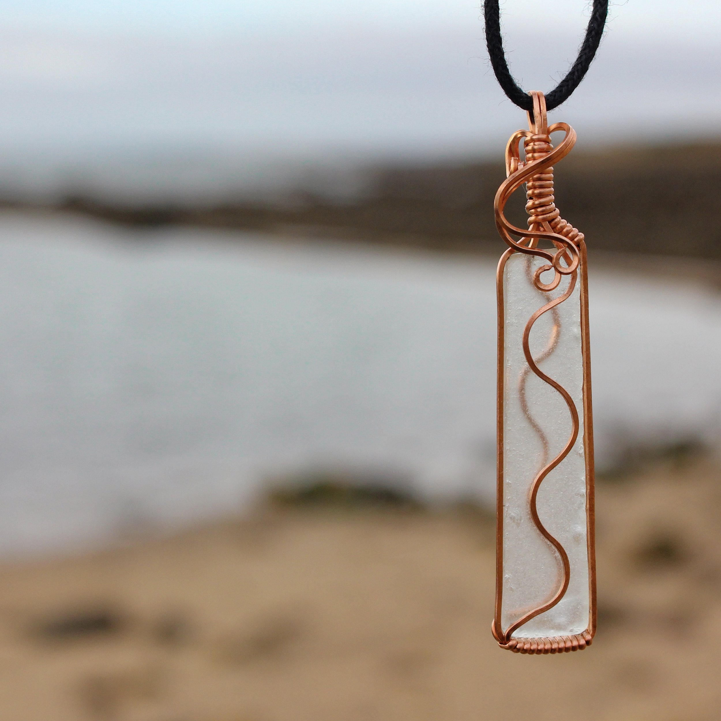 Seascape background with transparent sea glass pendant in foreground, the light depicts the copper wrapped swirls on both sides of the piece