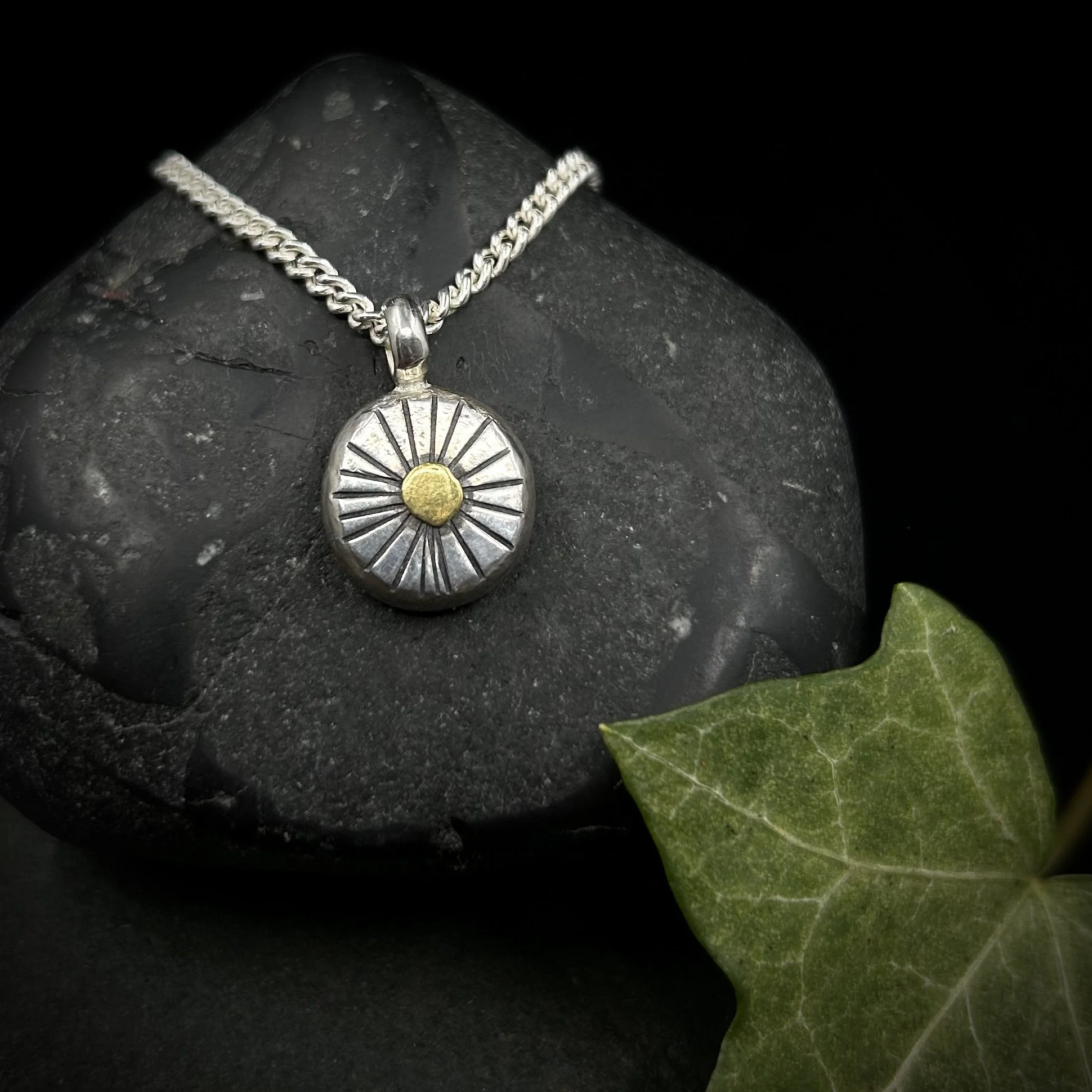 Solid silver circular pebble with textured lines radiating from a solid circle of 24k gold in the centre, set on a silver chain against a grey rock with green ivy beside it