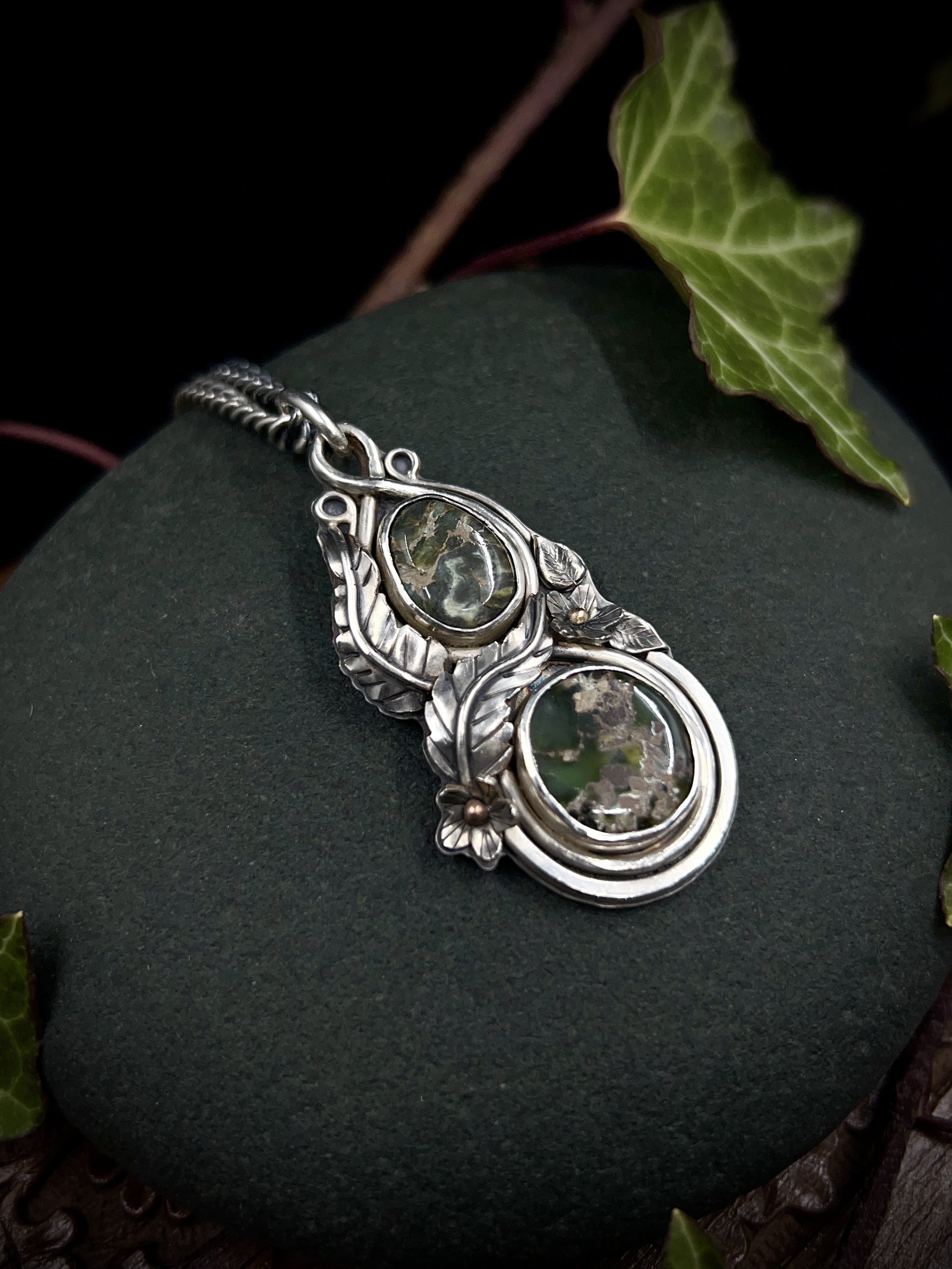 Large silver elven pendant with two green variscite stones and floral design elements on a grey rock with ivy