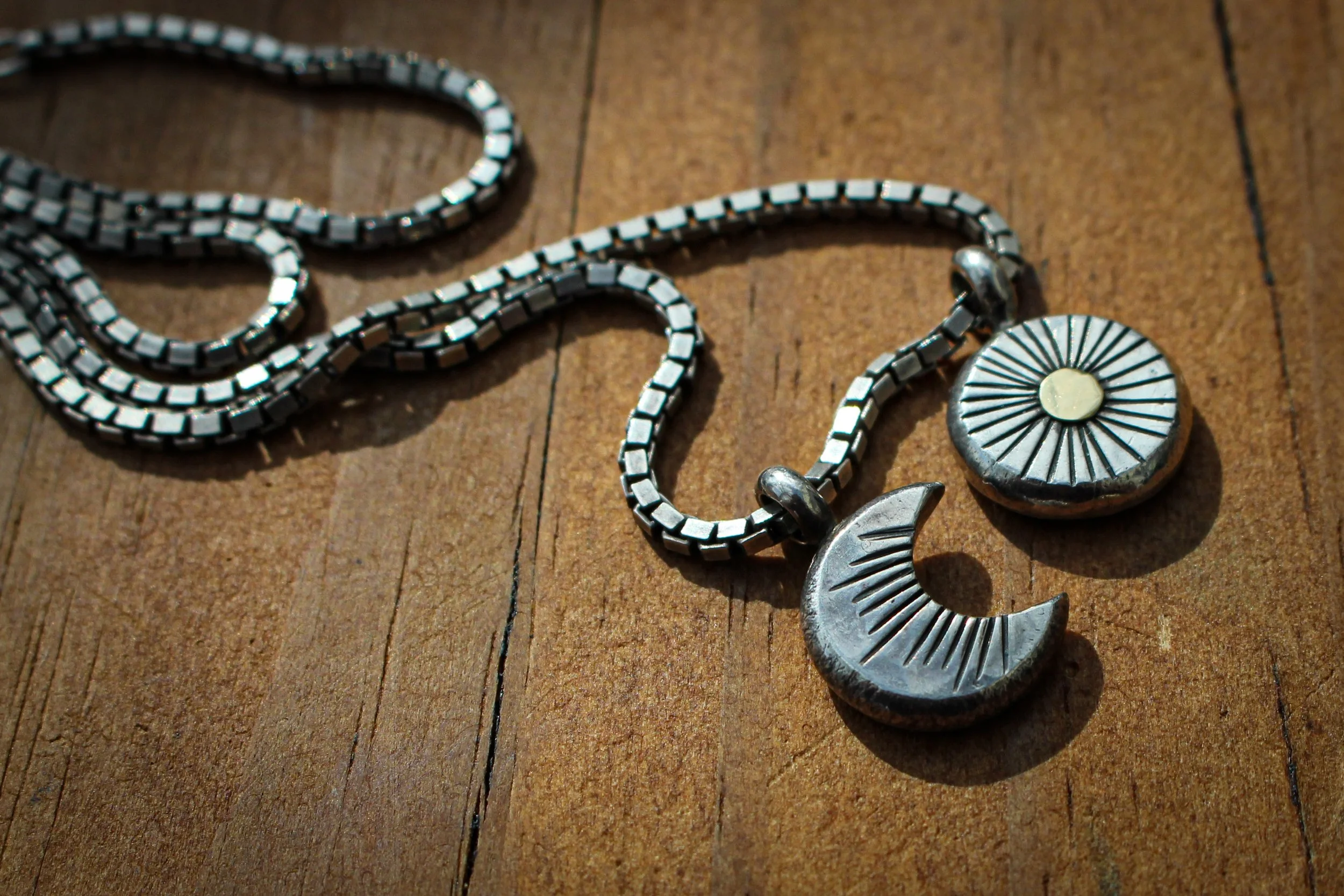 Two solid silver forged pendants, a crescent moon and sun with gold detail, are attached to a silver box chain lying on a wooden bench.
