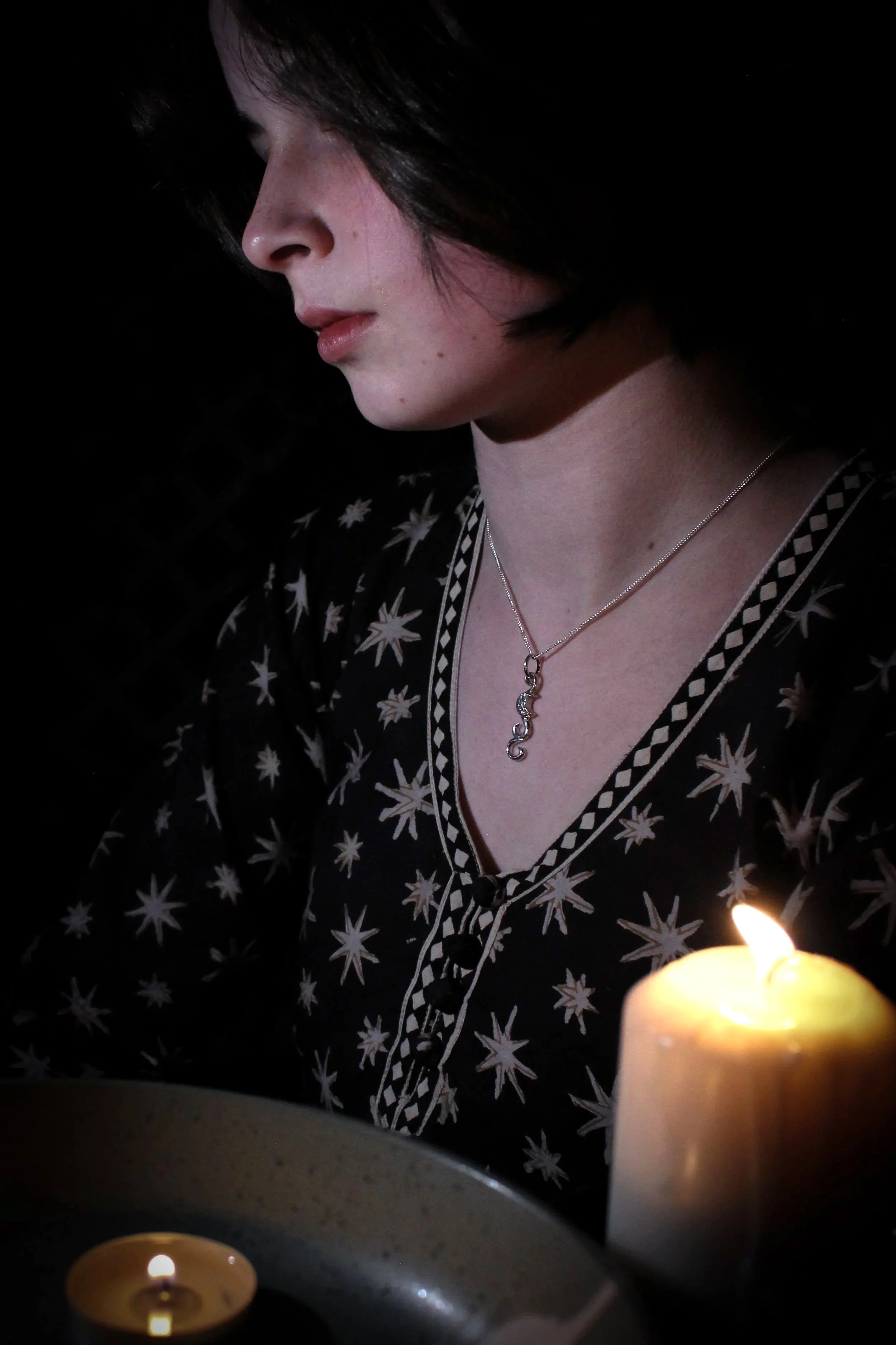Model wearing a black star print dress bathed in candlelight with a silver Moonglade pendant, crescent moon and swirls of silver, around her neck.