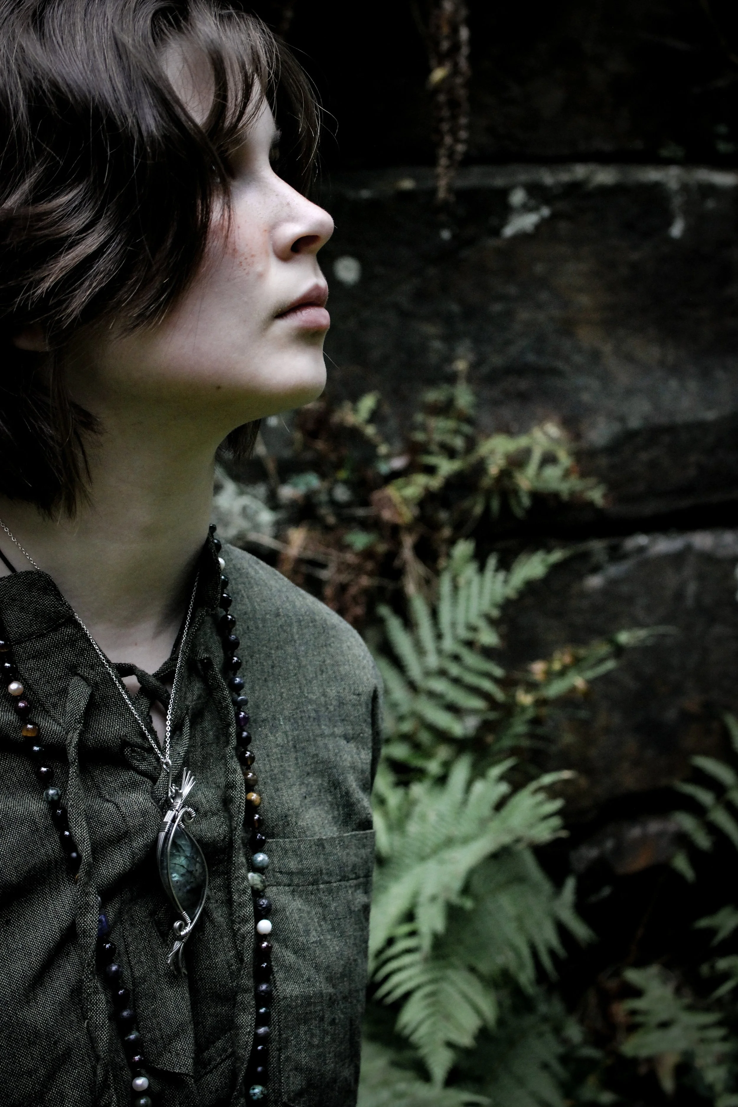 Model wearing a green shirt and a large labradorite and steel statement pendant with intricate construction and spiral design. Model is positioned beside an ancient wall with ferns