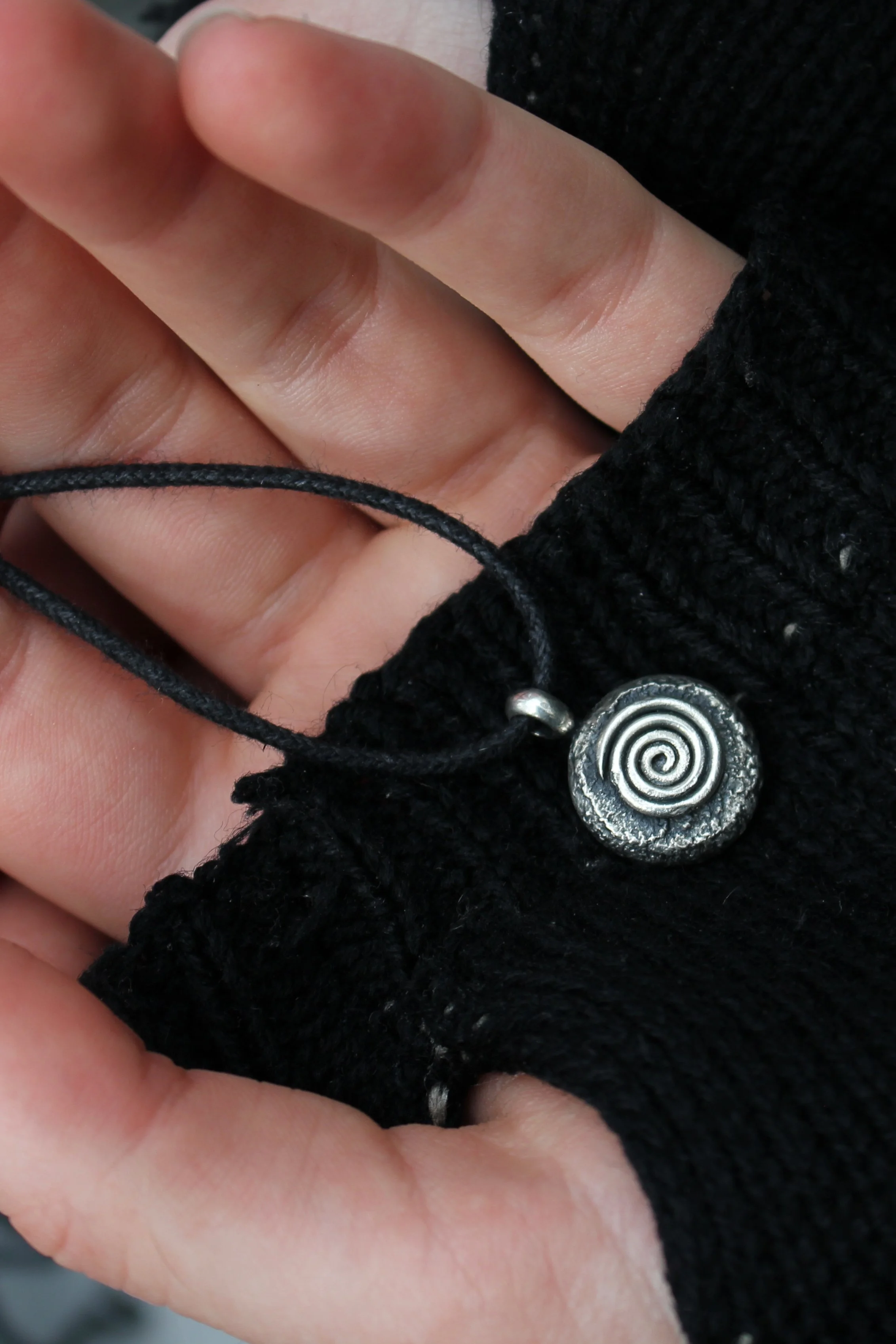 Round heavily textured and oxidised silver pendant with raised spiral on black cord laying on model’s palm