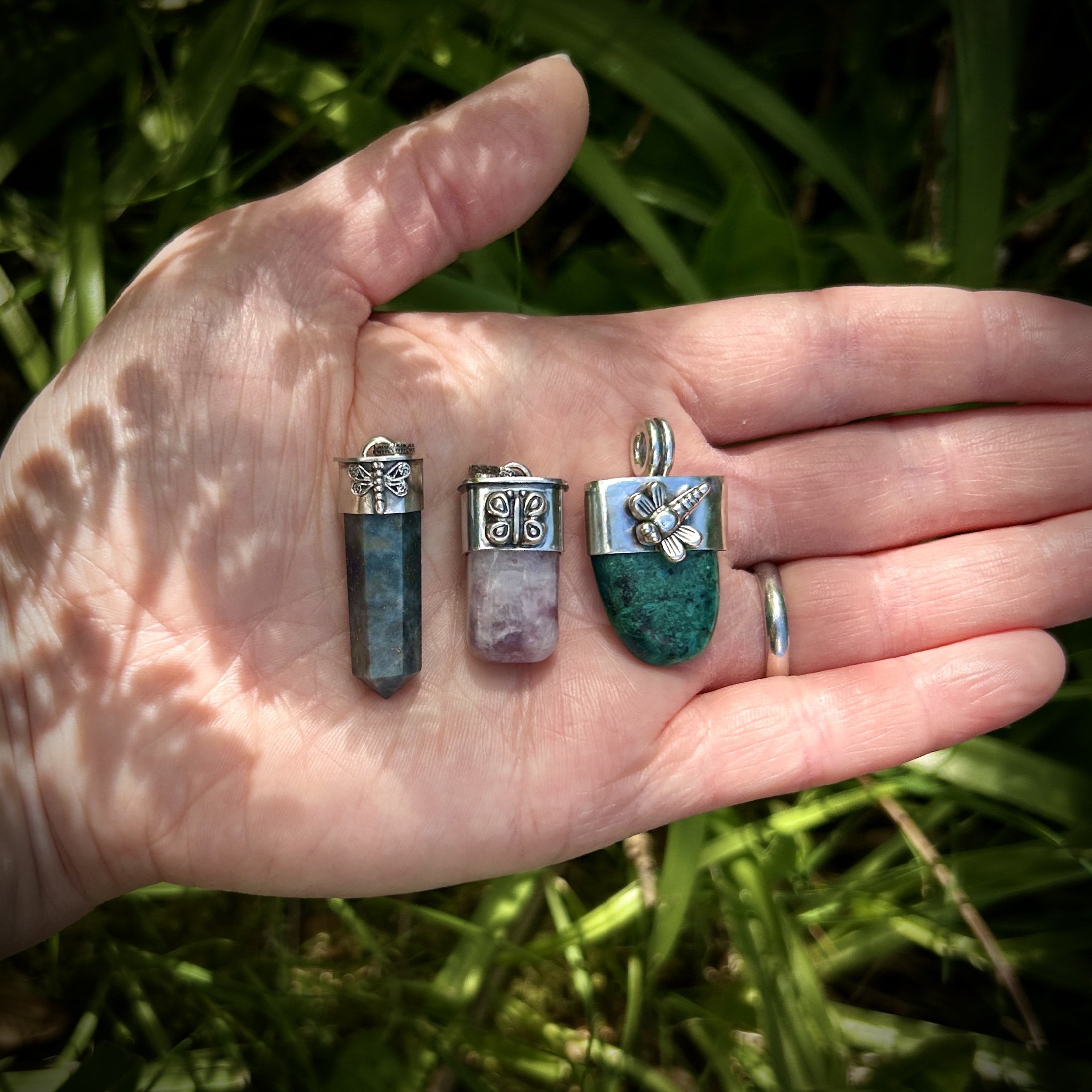 Scale photo in dappled sunlight of three crystal pendants with sterling silver butterfly and dragonfly motifs