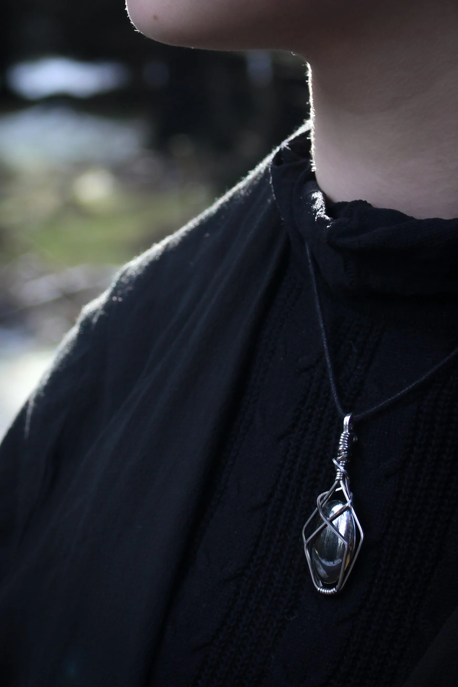 Close up of a model in a black top wearing an oval hematite pendant, the reflective stone is wrapped in steel in such a way as to resemble a shield with a cross