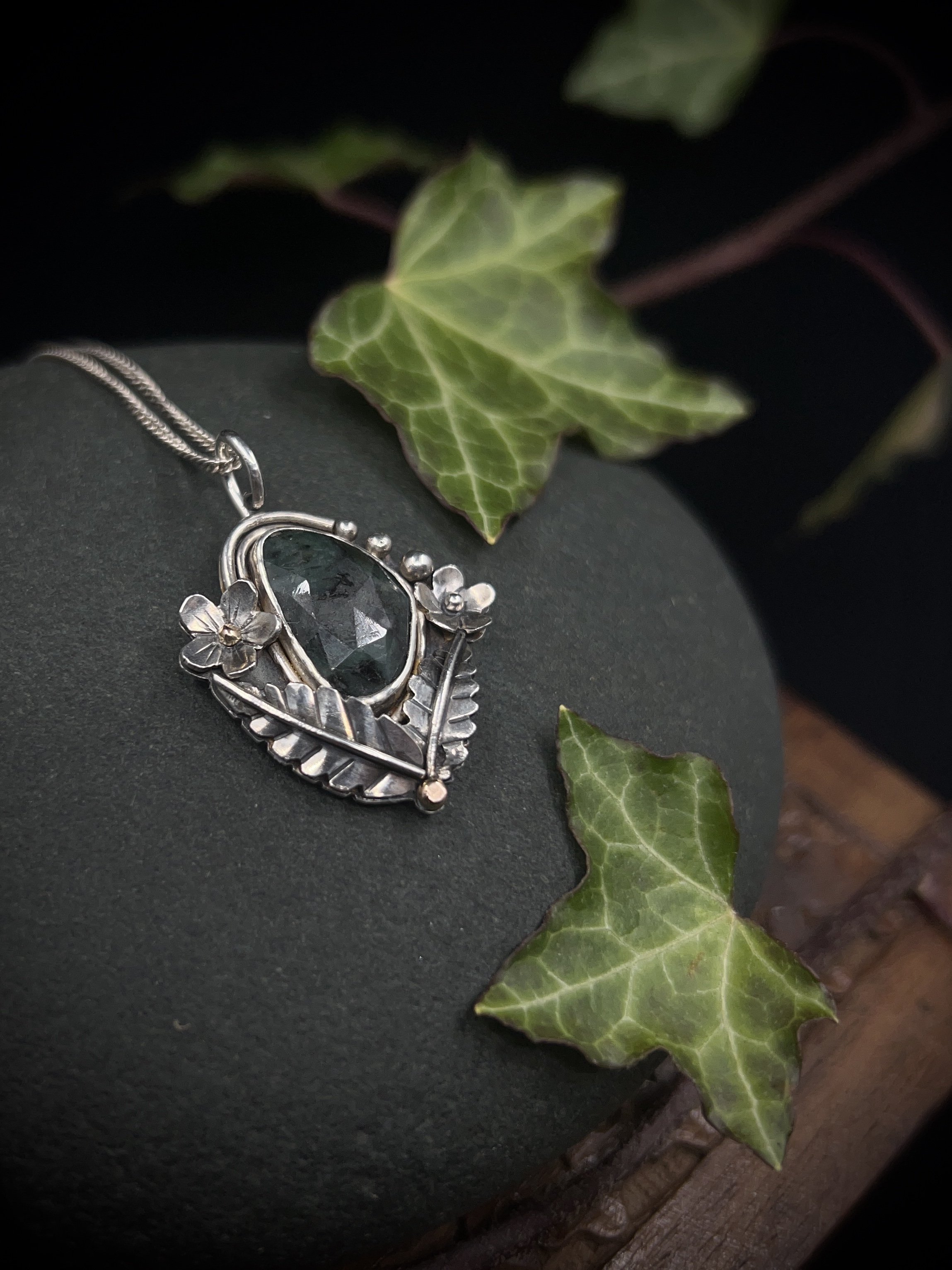 Silver floral pendant with a raw faceted dark green emerald in the centre surrounded by leaves, flowers, buds and rose gold details