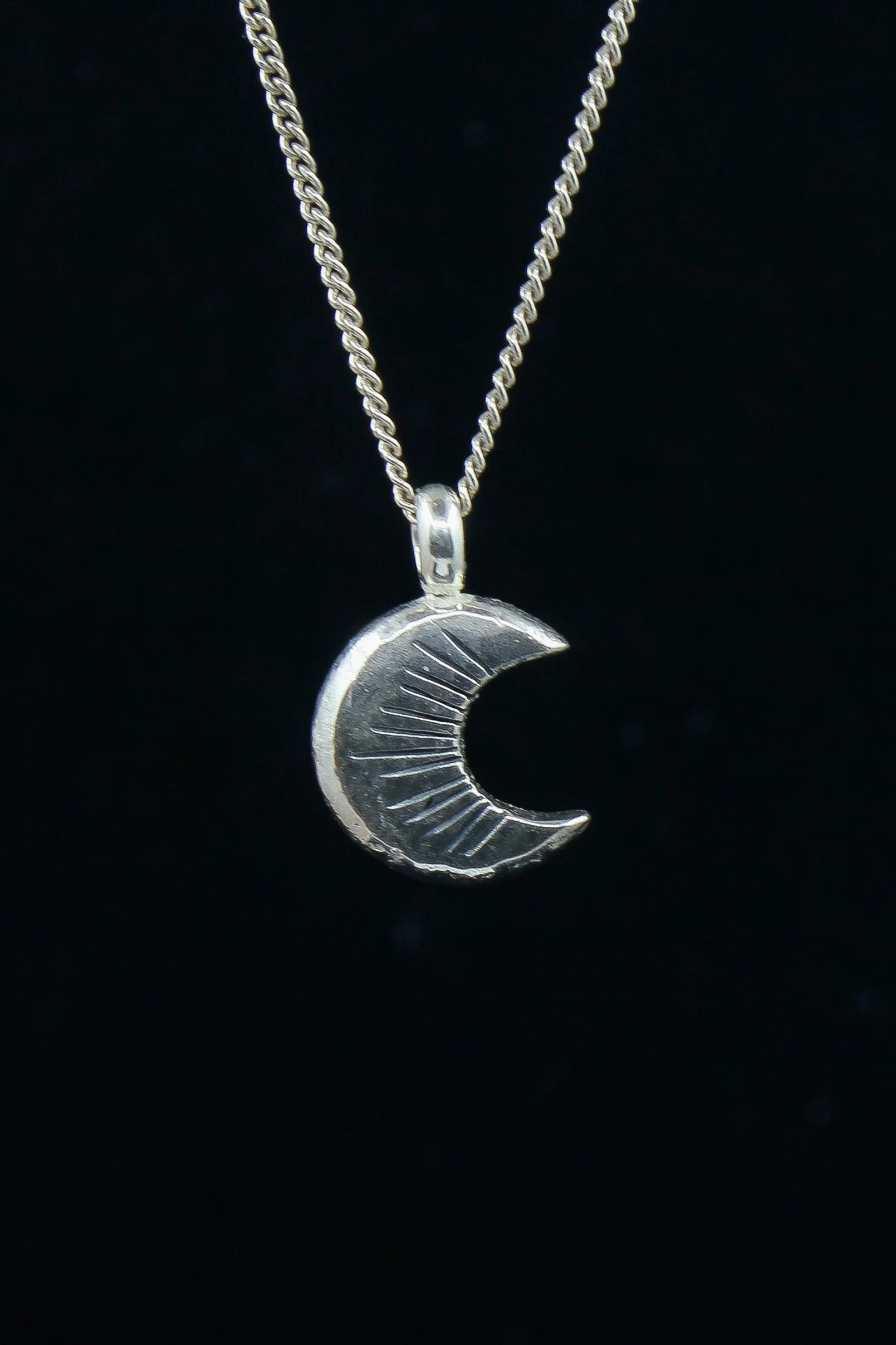 A silver forged crescent moon hanging from a silver chain against a black background. The moon pendant displays thirteen oxidised notches.