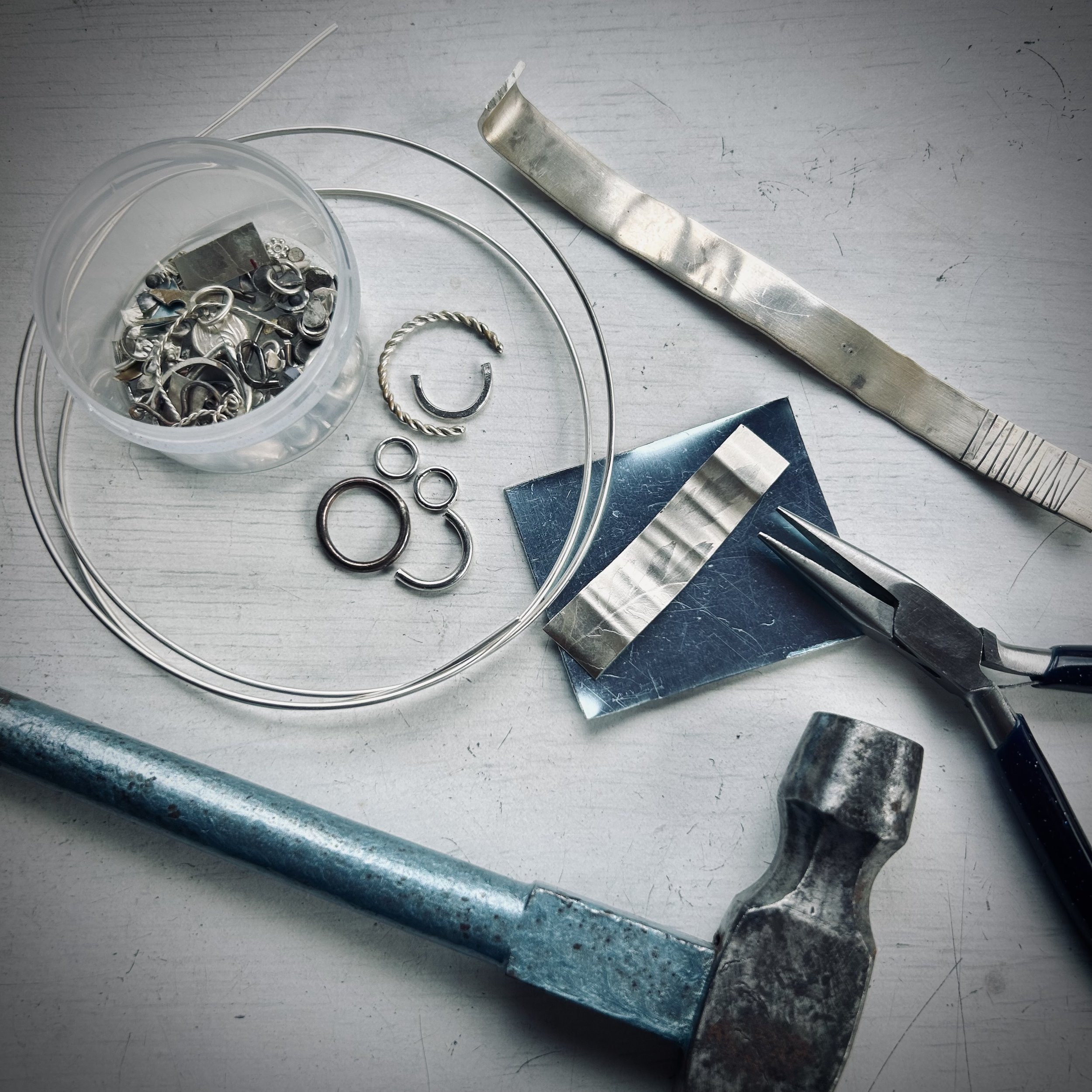 A selection of sterling silver - such as sheet, wire, jump rings and other bits and pieces - laid out with a pair of pliers and a hammer.