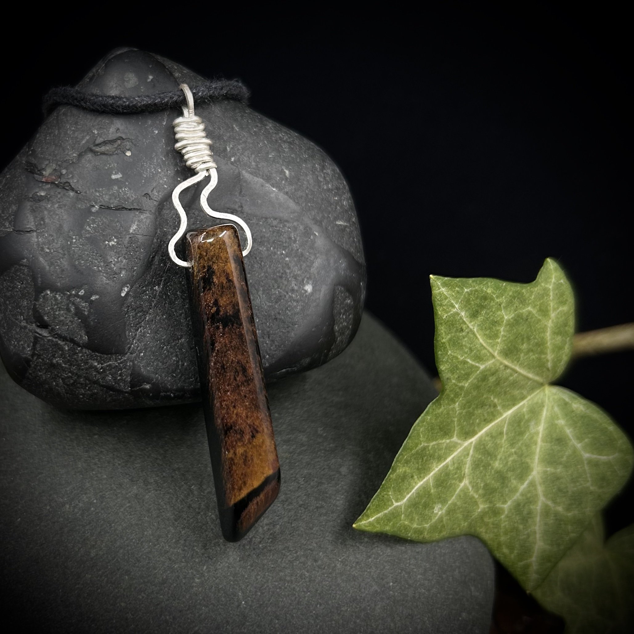Long brown tiger’s eye stone with a silver flame shaped wrapped top hanging from a black cord beside some ivy leaves