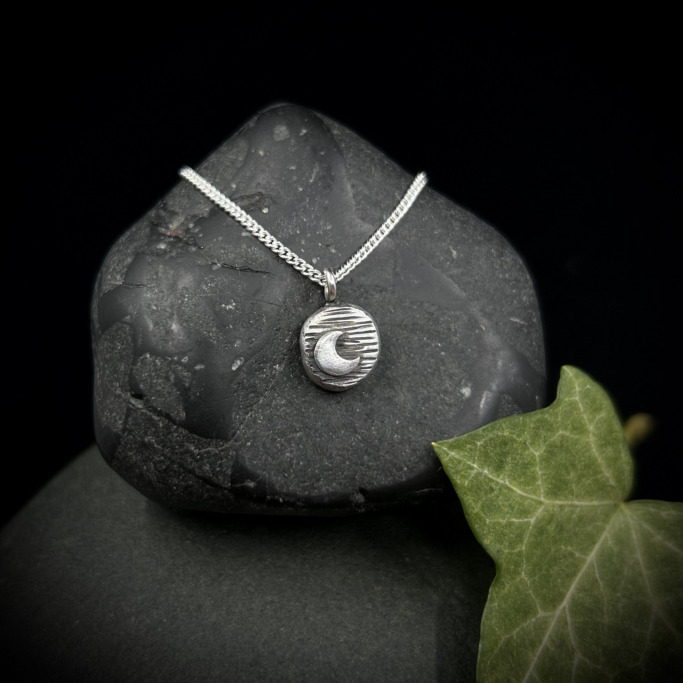 Silver crescent moon on a circular pebble of solid silver, the moon is emphasised by the darkened lights marked on the surface of the silver