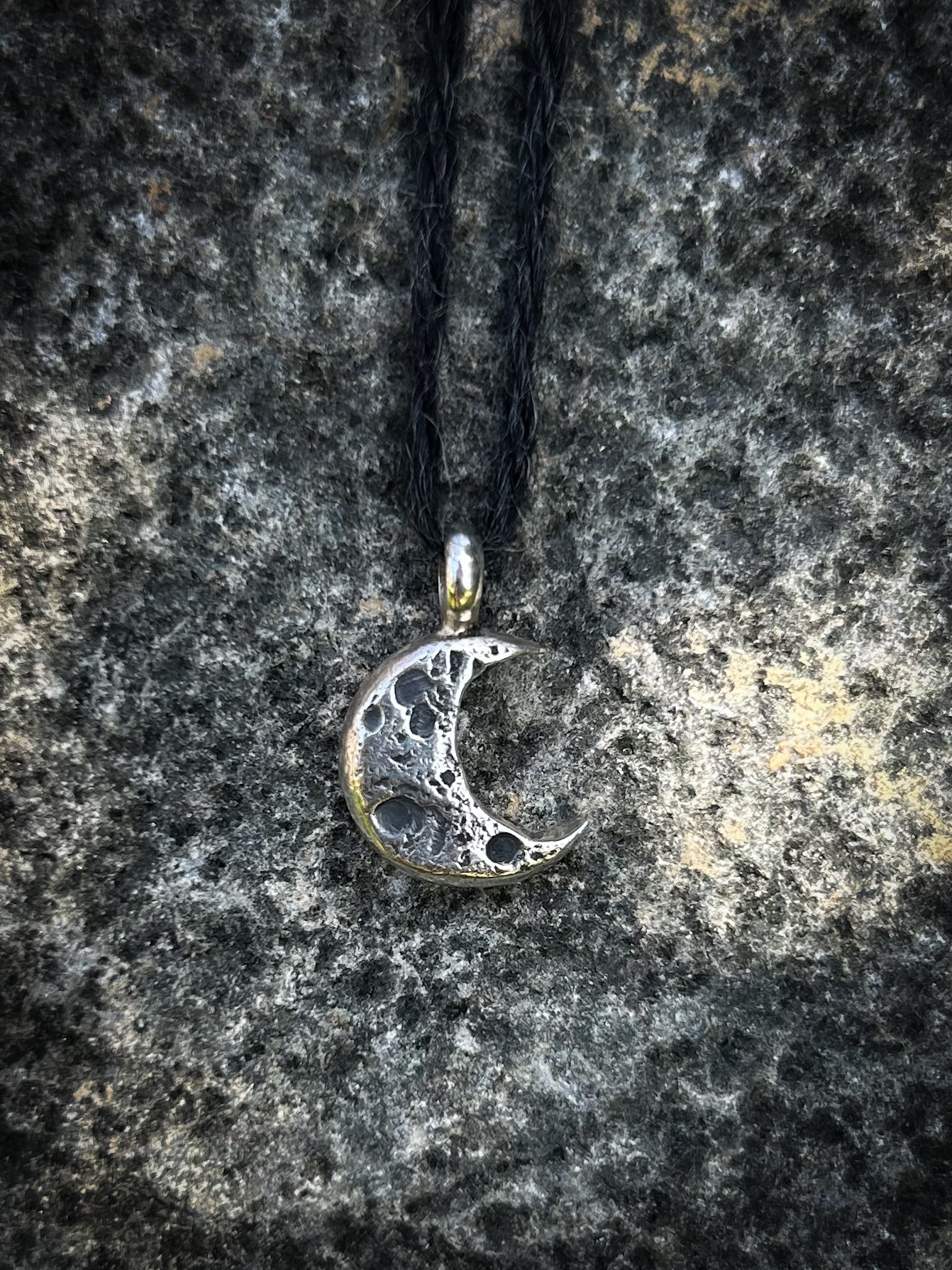 A textural silver crescent moon pendant with deep oxidised markings resembling the surface of the moon. The jewellery is suspended from black cord against a textured rock surface. 